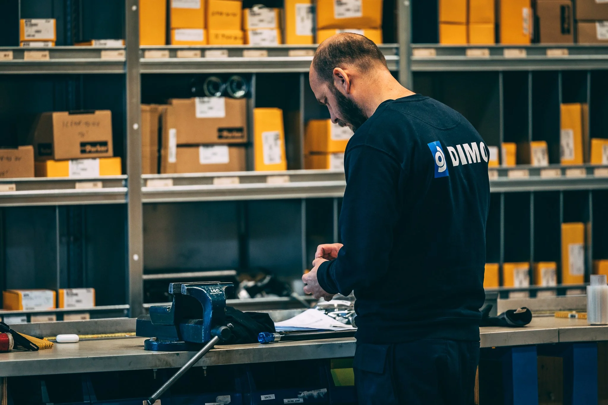 Slangeverkstad Hogne. HoseDoctor, Parker Store. En mann med kort hår og skjegg jobber på et lager, iført en svart DIMO T-skjorte, omgitt av hyller fylt med skjulte handleposer.