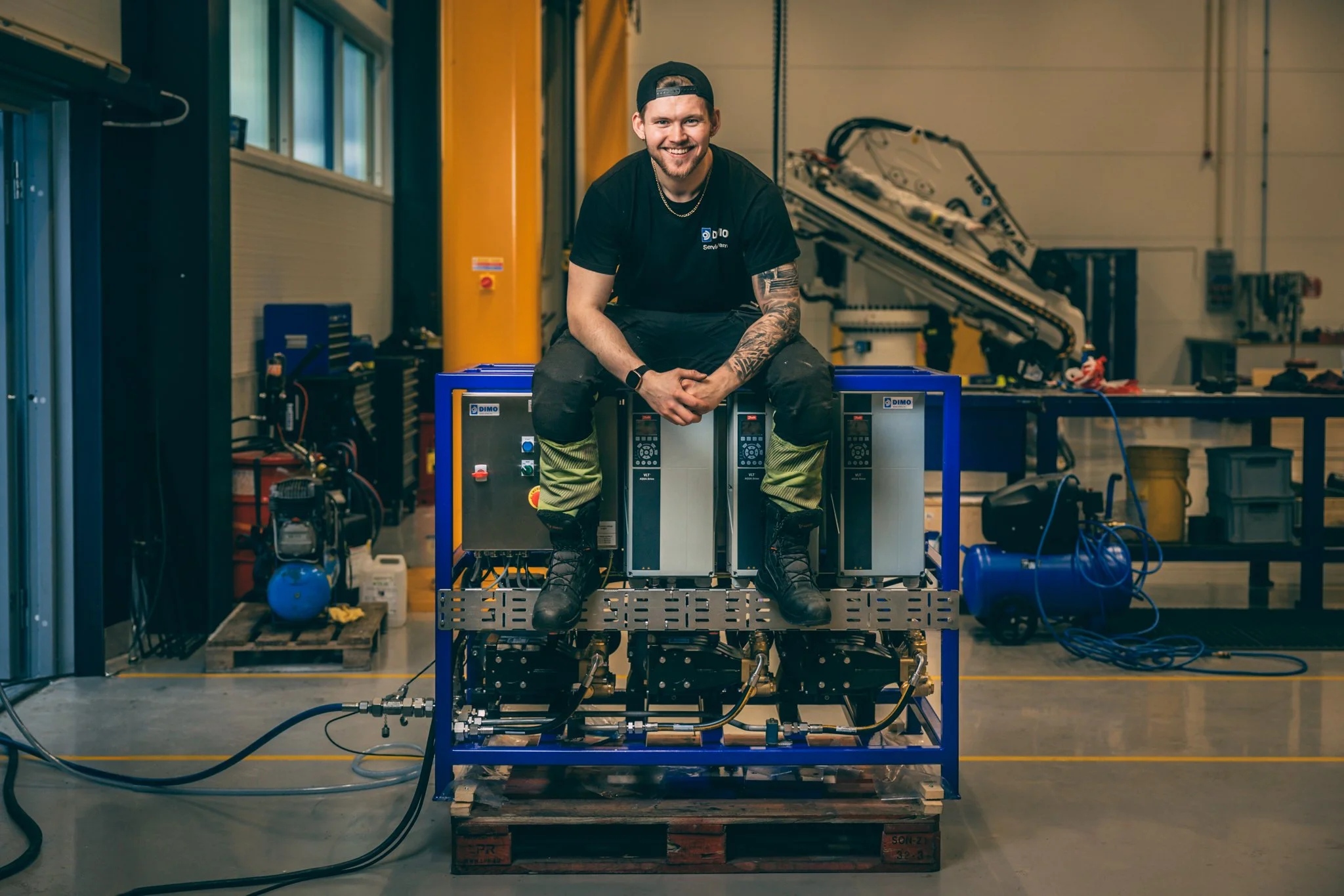 Dimo som lærebedrift, lærlinger og fagbrev. Maskinist, elektro, automasjon, mekaniker, produksjon, industri. En smilende mann i arbeidstøy i et verksted med maskiner og utstyr rundt ham.