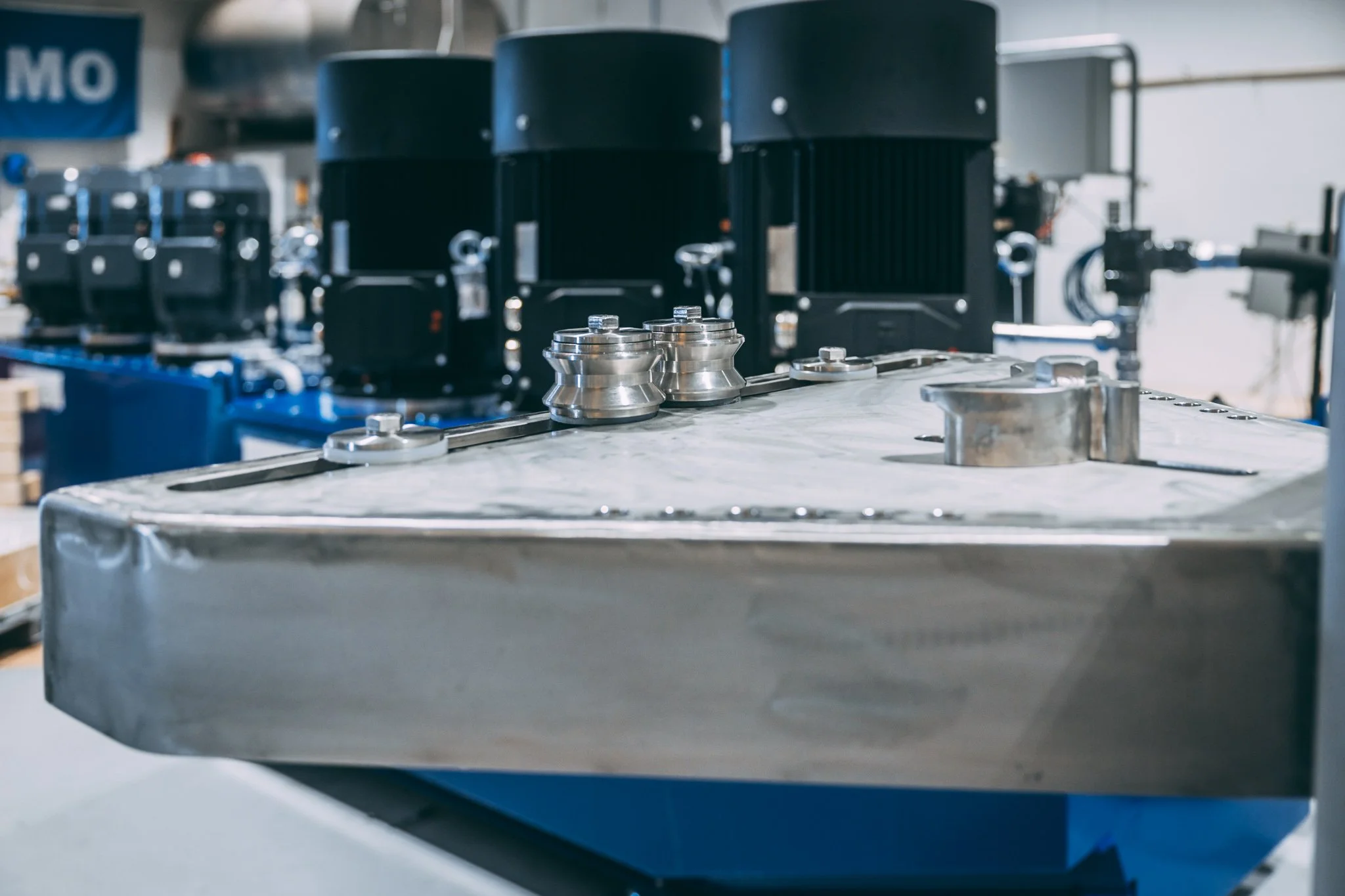 Bildet viser industrielle maskindeler på et verksted eller fabrikk, inkludert metallkomponenter og store svarte maskiner i bakgrunnen.