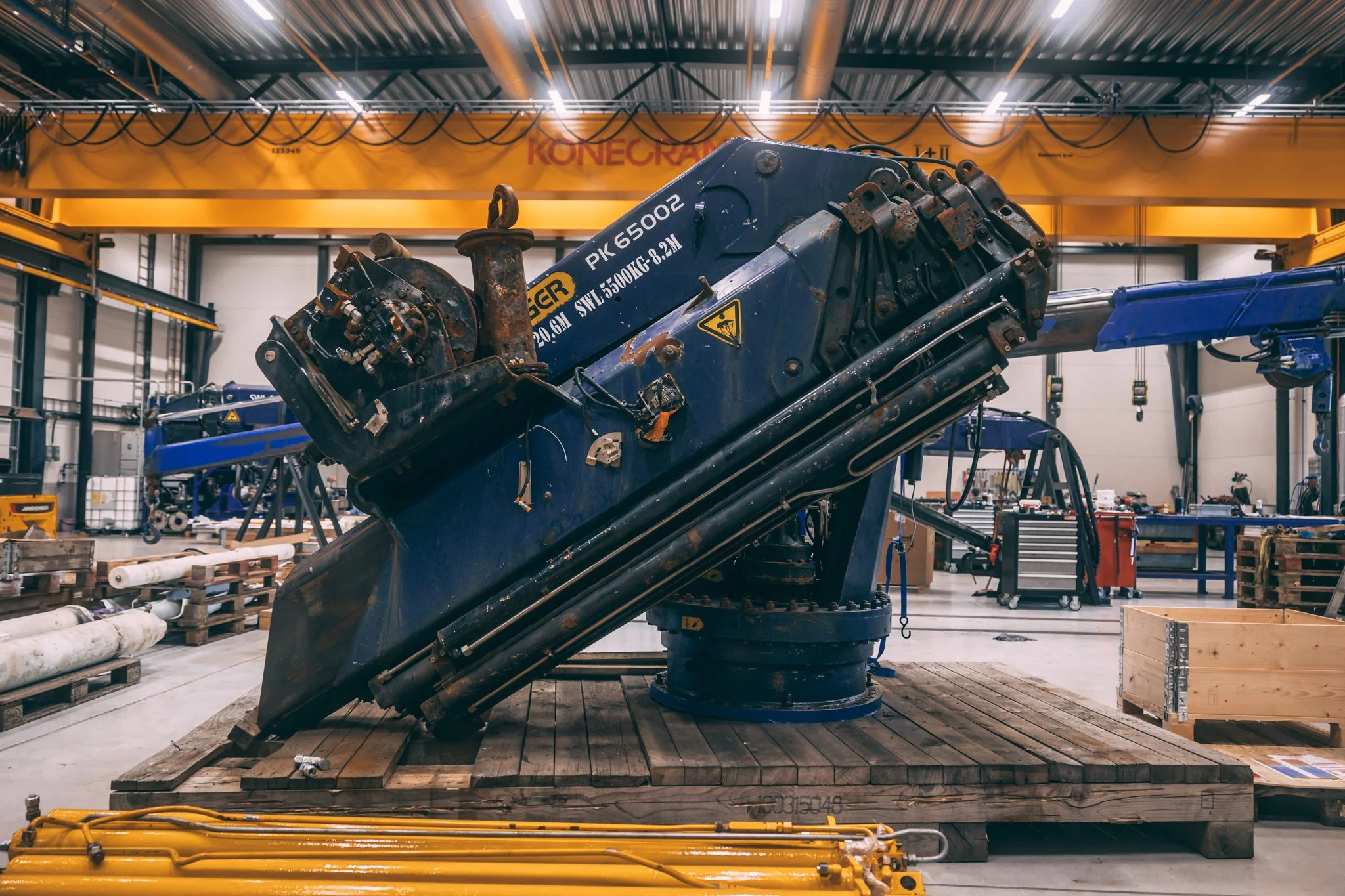 Dimo har kran inspeksjon. Industrielt verksted med stor kran på opphøyde planker, omgitt av arbeidsutstyr, verktøy og stålbjelker. HS marine.
