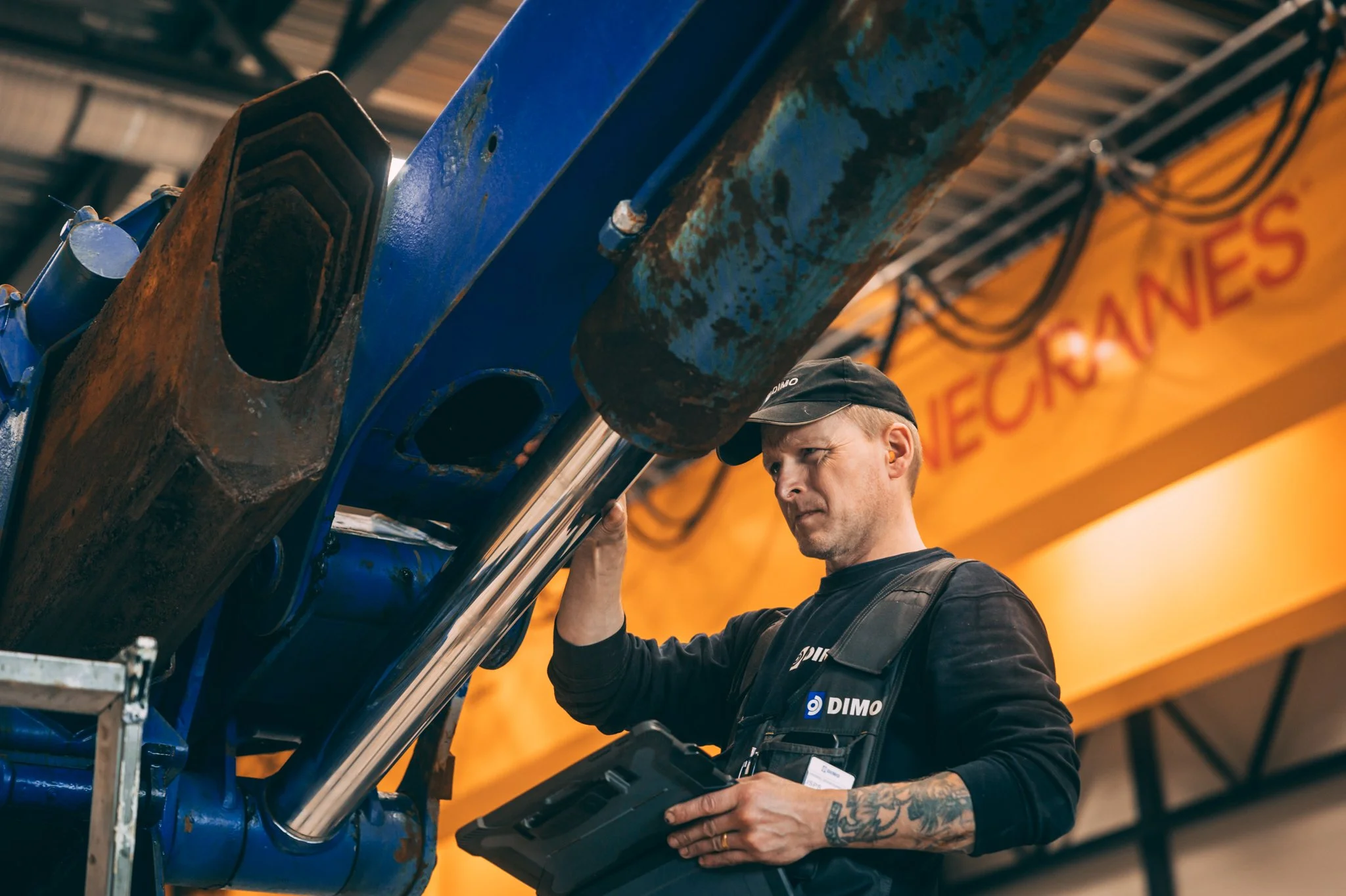 En mann med tatoveringer på armen jobber med en stor blå maskin i en industriell setting. Mannen bruker en lue og holder et kontrollapparat.