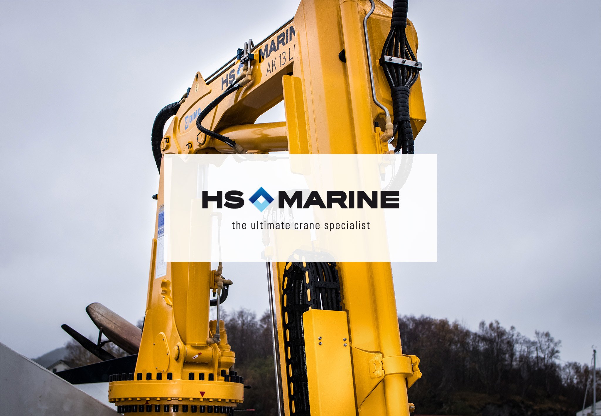 En stor, gul kran med logoen til HS MARINE og tekst "the ultimate crane specialist", satt opp utendørs med en overskyet himmel bak.