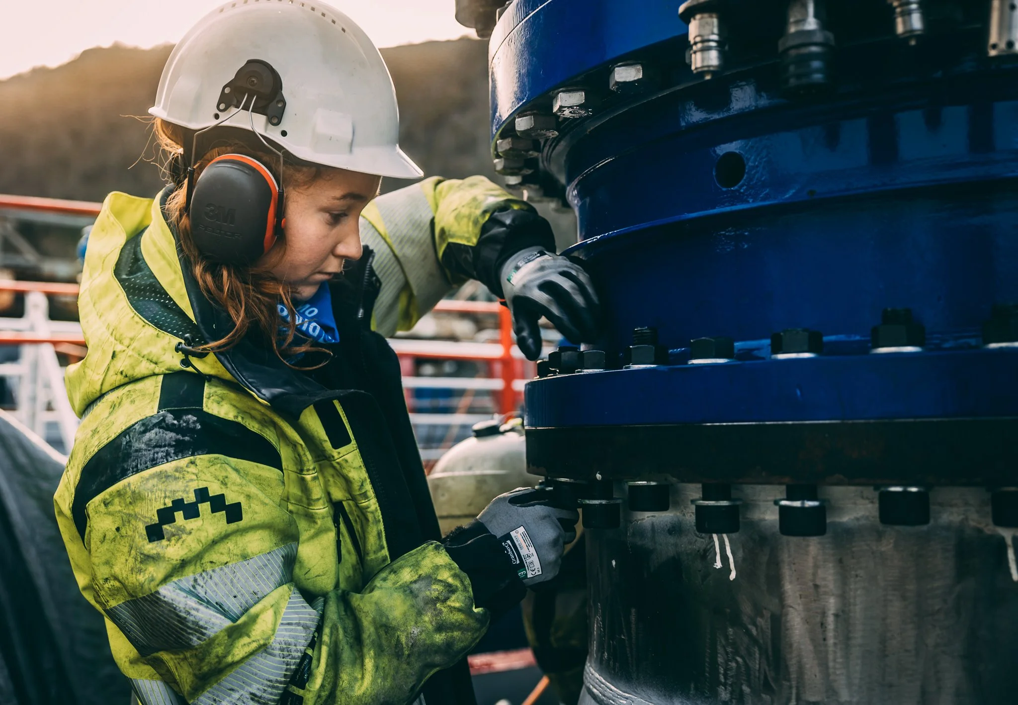 Palfinger, Dimo Regina. HMS. En kvinne i gult arbeidsantrekk og hjelm jobber med mekanisk utstyr, i en industriell og maritim setting. En kvinne i sikkerhetsutstyr inspiserer en stor maskindel utendørs under solnedgang. visibility work wear.