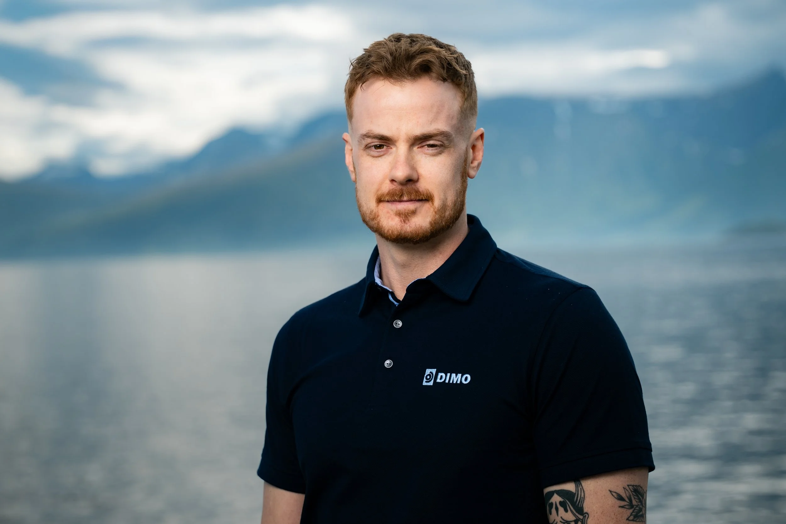 Ung mann med lettfrettet hår og skjegg i mørk DIMO t-skjorte, står utenfor med en fjell- og vannbakgrunn.