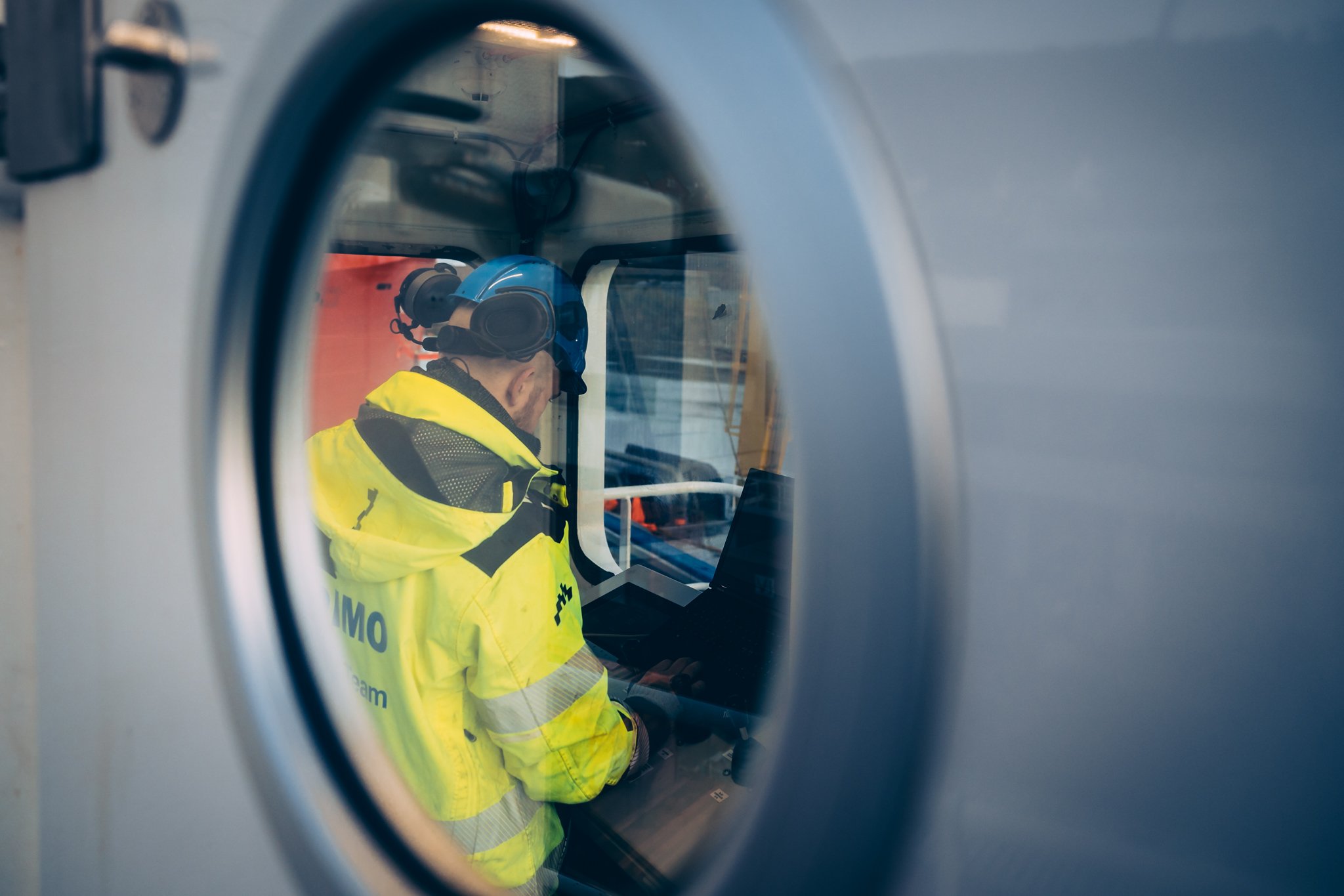 IT infinity Vard. Dimo arbeider iført gul jakke, high visibility work wear, arbeidskle, og hjelm, sett fra eit vindu i dør, som ser inn mens personen jobber.