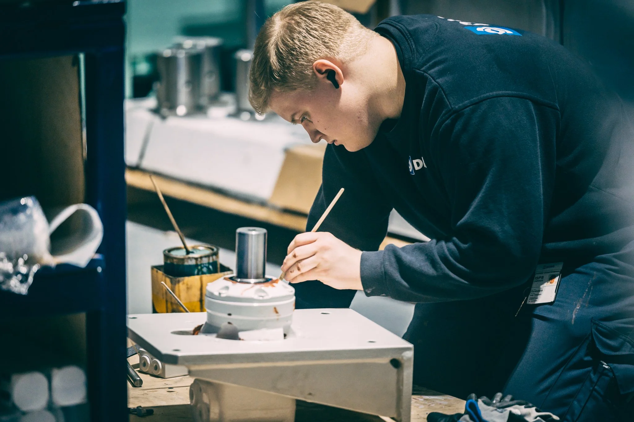 En ung mann med blond hår, iført svart genser, jobber med et teknisk prosjekt på et arbeidsbord i et verksted med en vinsj, pal winch.