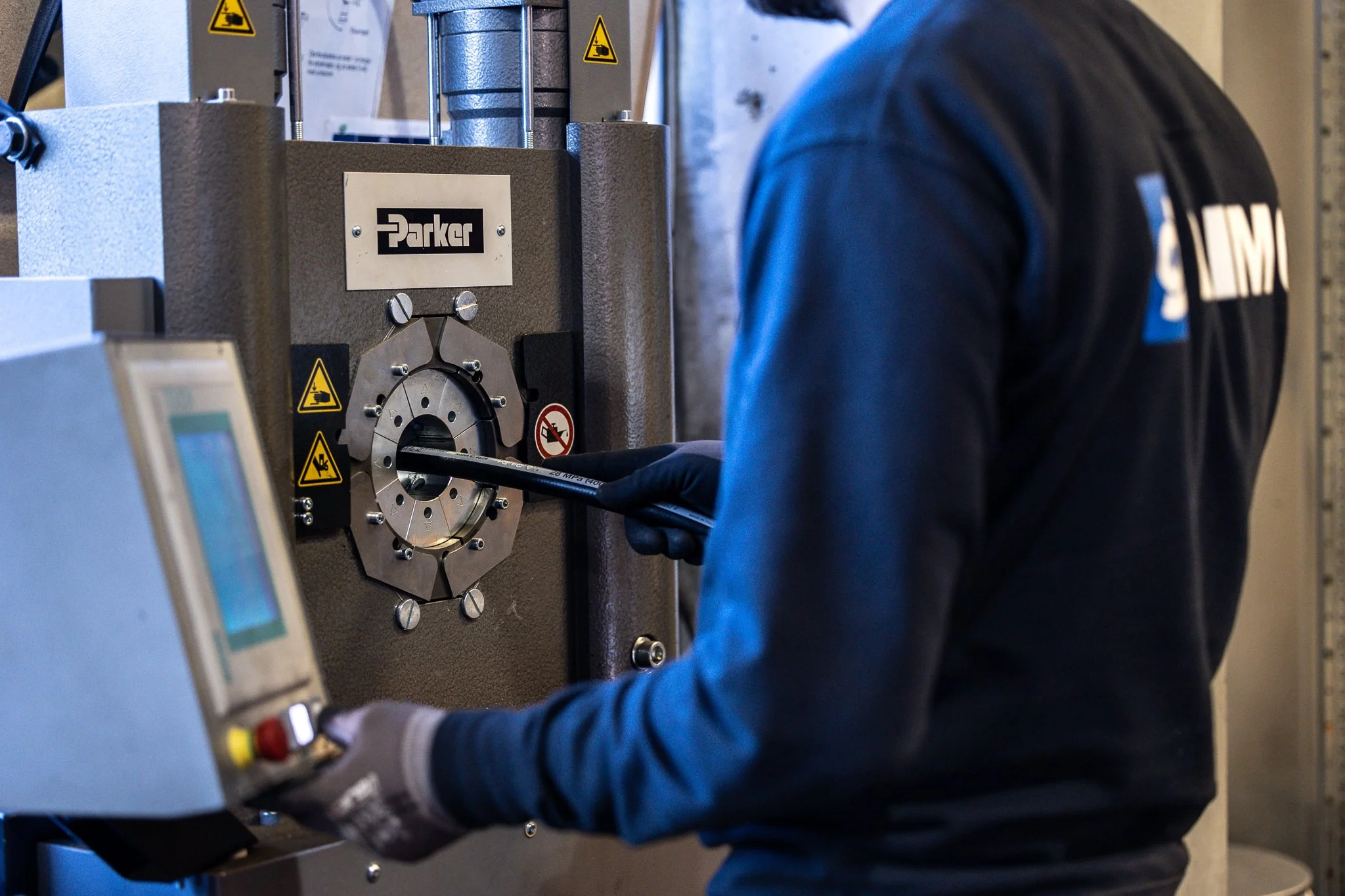 HoseDoctor - parker sertifisert. Dimo - En person i blå arbeidsklær jobber med maskineri merket Parker. Det er synlige advarselsskilt på maskinen.