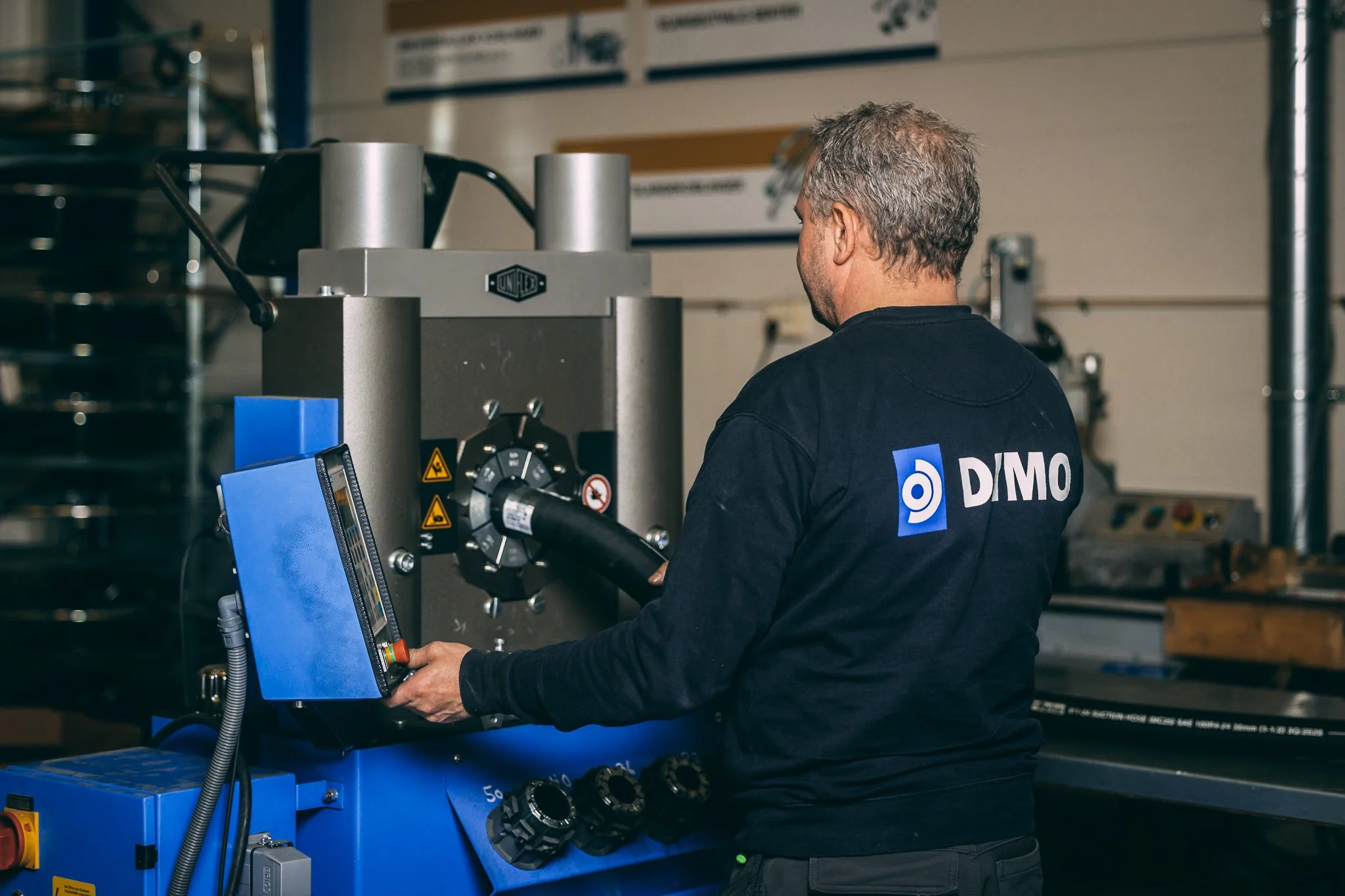 En mann i svart T-skjorte med DIMO-logo jobber med maskineri i et industriområde.