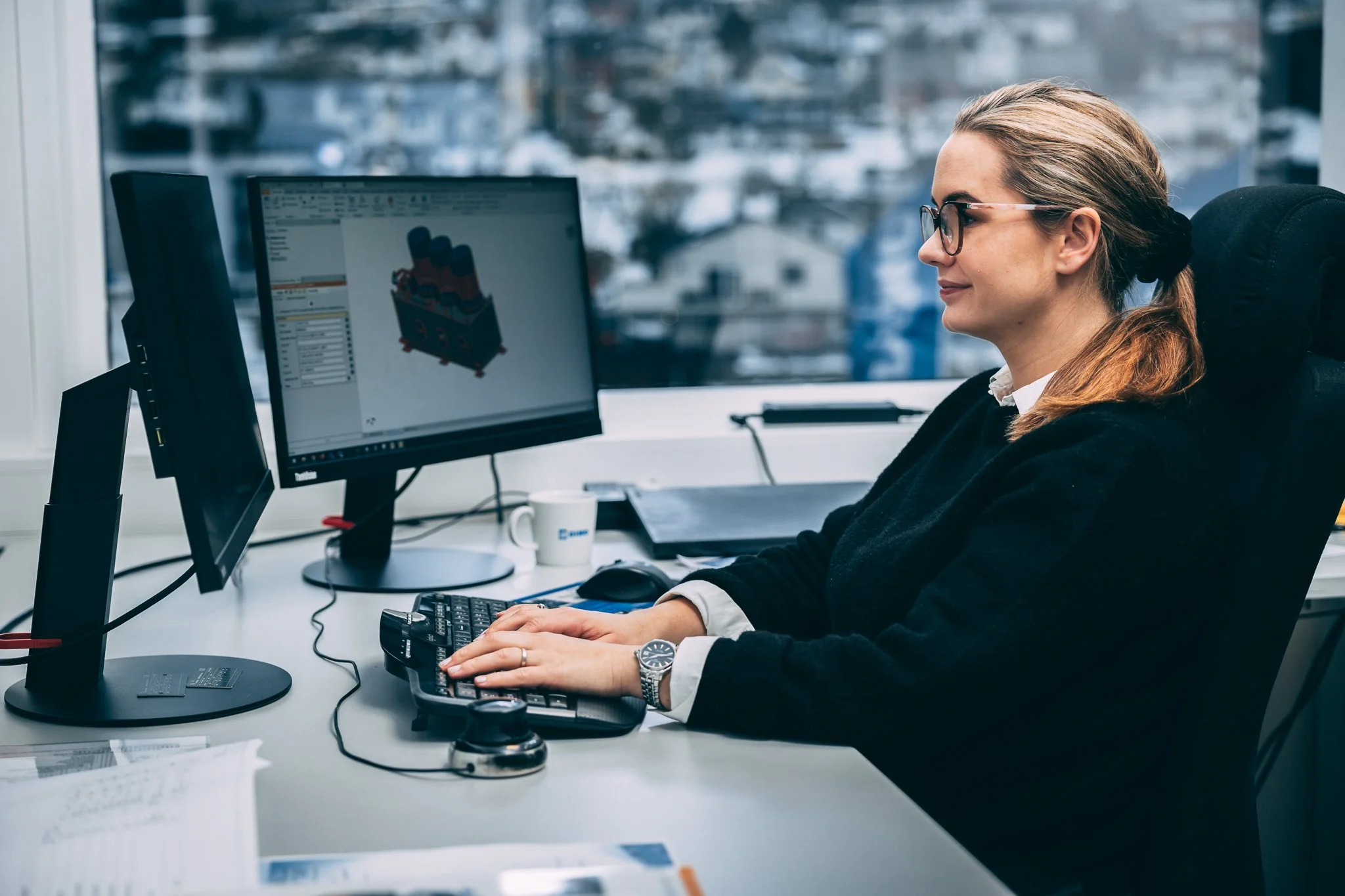 Kvinne som jobber på kontor, skriver på tastatur, med to skjermer og papirer på skrivebordet, utsikt over by utenfor vindu.