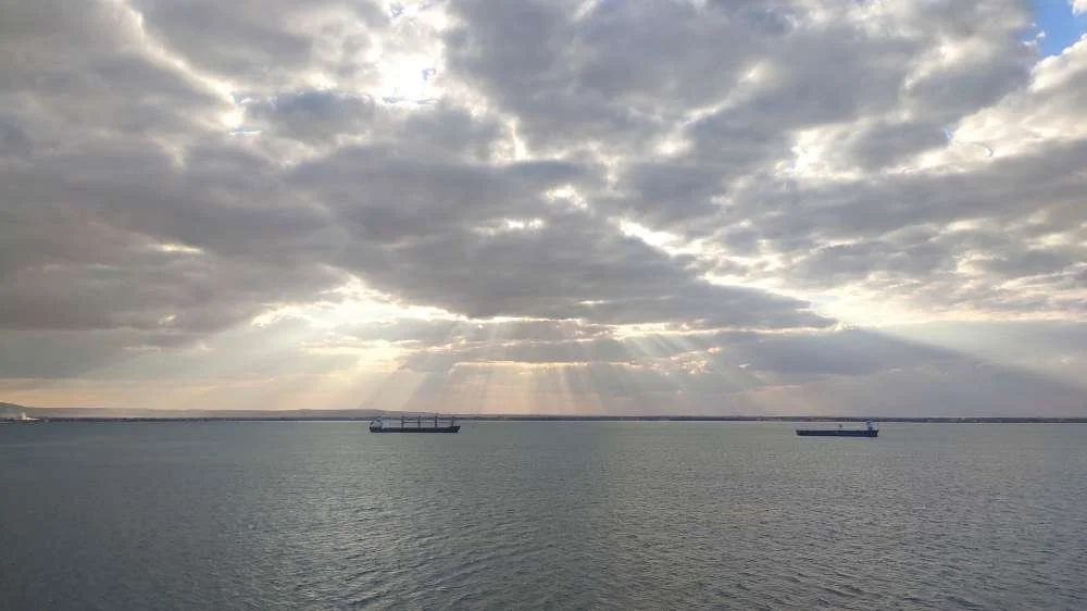 A body of water with two ships floating, and a cloudy sky with rays of sunlight breaking through.