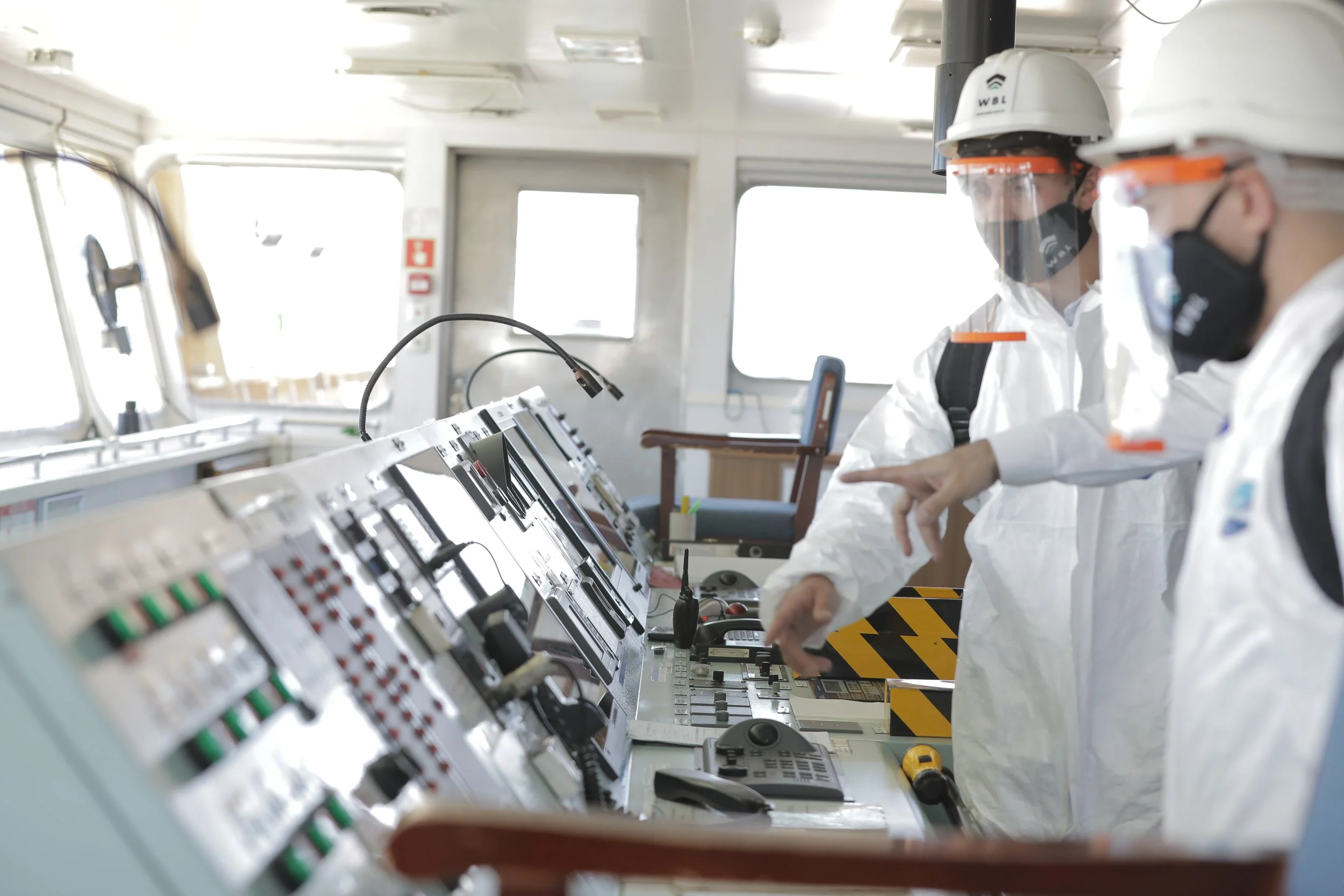 Workers in protective gear, including helmets and face masks, operating control panels on a ship's bridge.