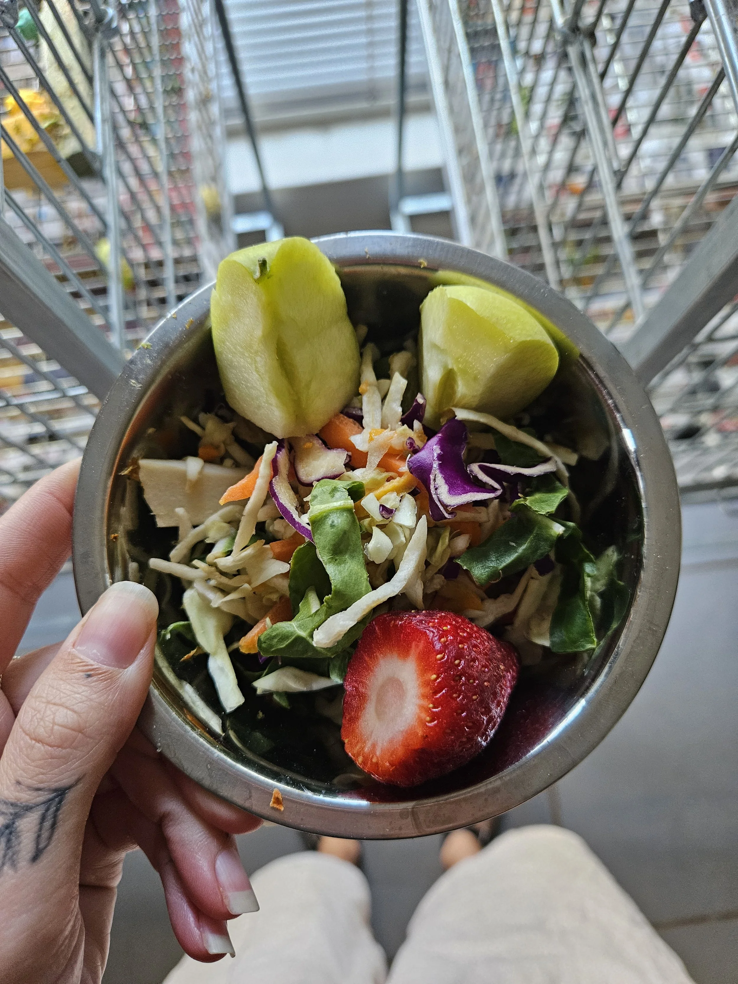Close-up of a salad in a metal bowl with green apple slices, shredded cabbage, and a strawberry, held by a person with painted nails in front of a shopping cart.