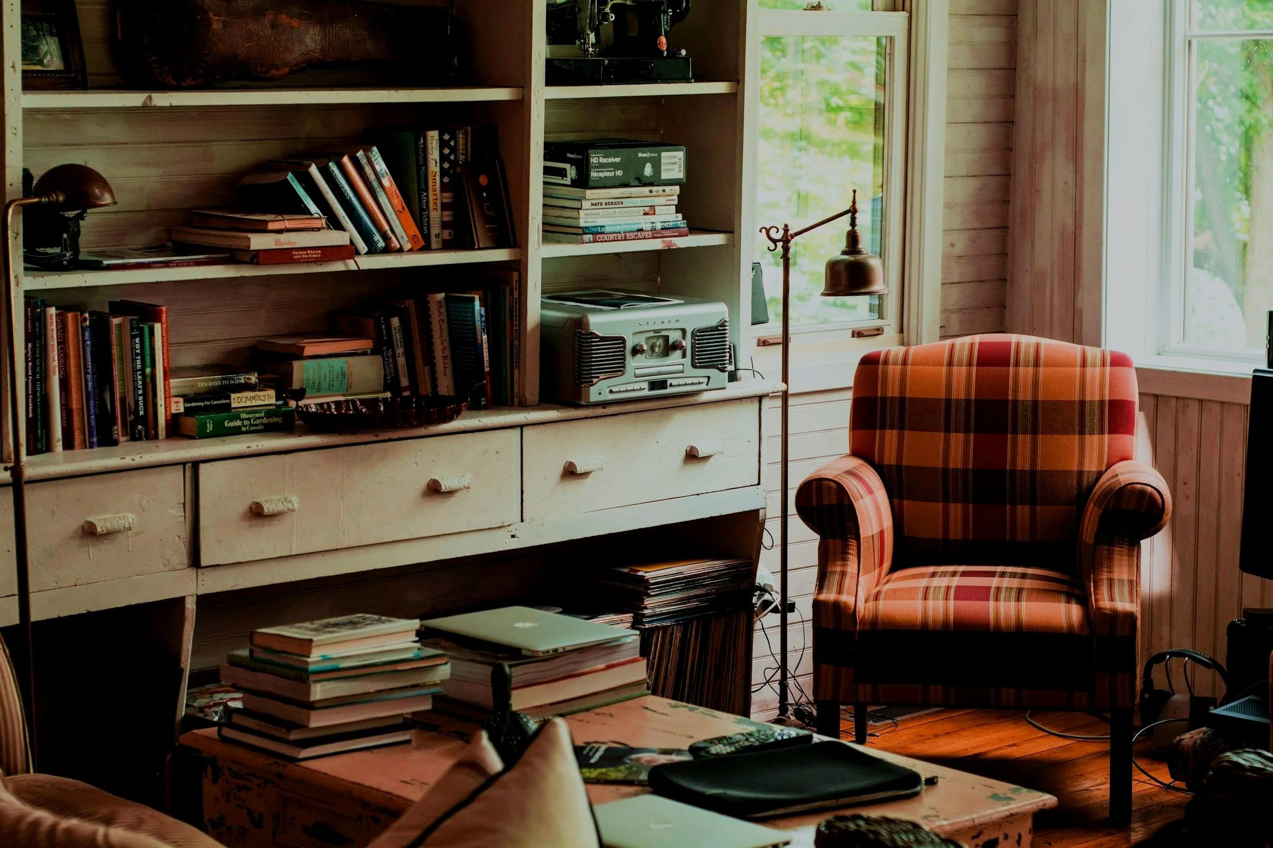 A cozy room with a wooden bookshelf filled with books and some electronic equipment. There is a plaid armchair, a wooden table with books, and a window showing green trees outside.