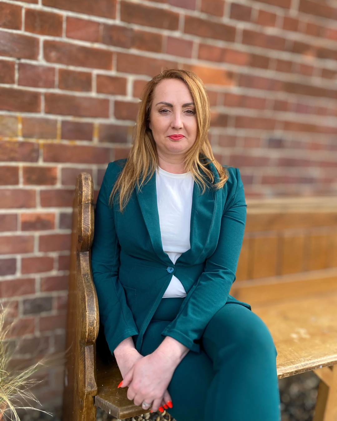 A woman with red hair and fair skin sitting on a wooden bench against a brick wall. She is wearing a teal blazer and matching pants, and a white shirt. She has bright red lipstick and nail polish, and looks directly at the camera with a neutral expression.