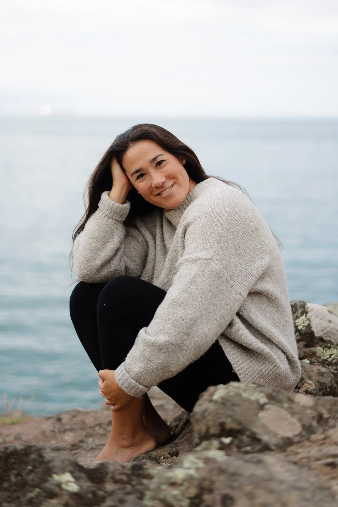 A woman sitting on rocks near the water, smiling and wearing a beige sweater and black pants.