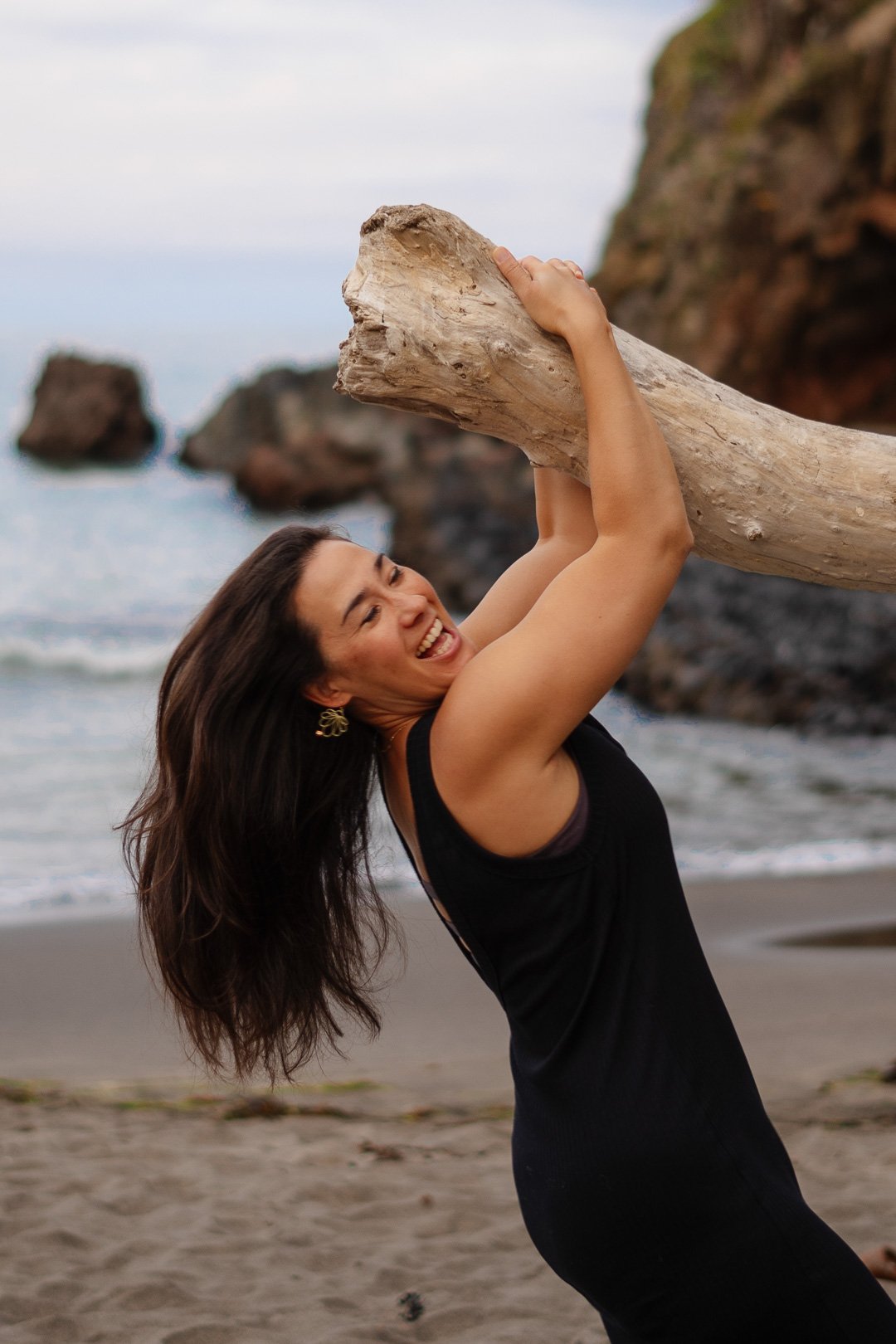 Woman with long dark hair smiling and holding a large piece of driftwood on a beach with rocks and the ocean in the background.