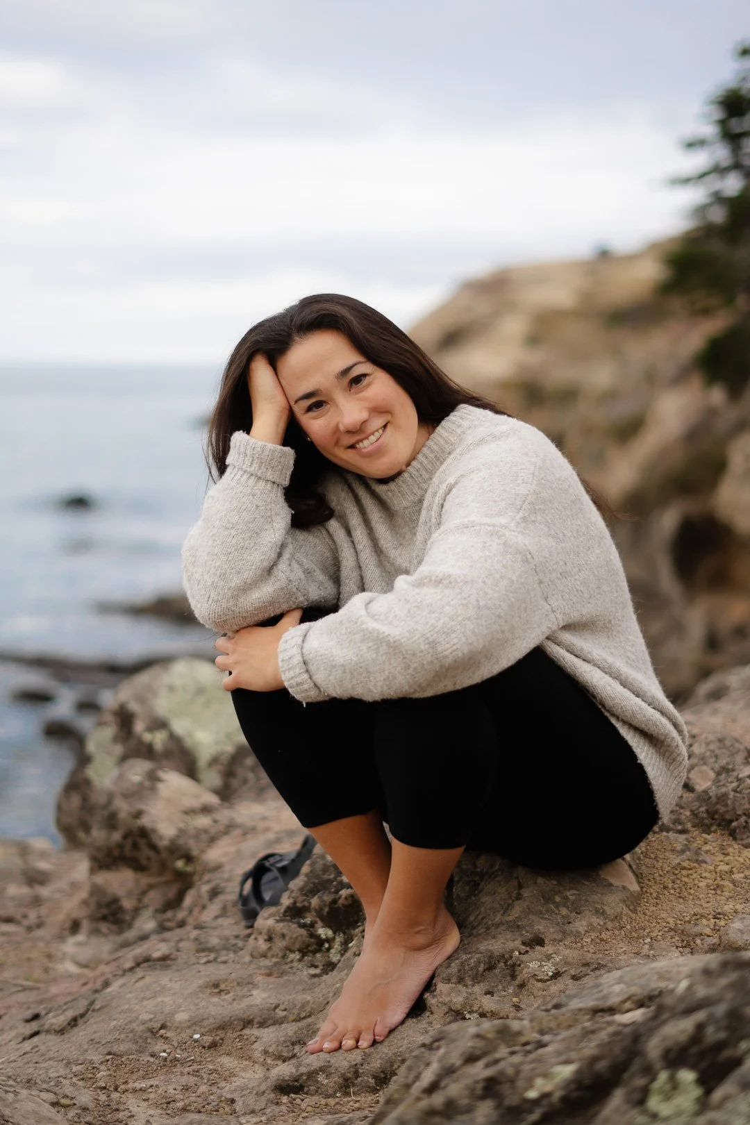 A woman with long dark hair, sitting on rocks near the shore, smiling at the camera. She wears a beige sweater and black pants, with a backdrop of water and rocky cliffs.