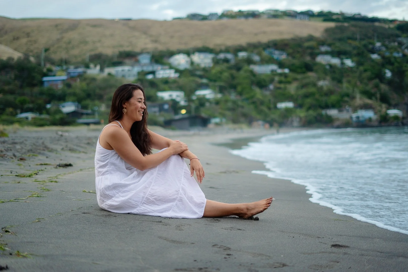 Woman in a white dress sitting on the beach near the water with green hills and houses in the background.