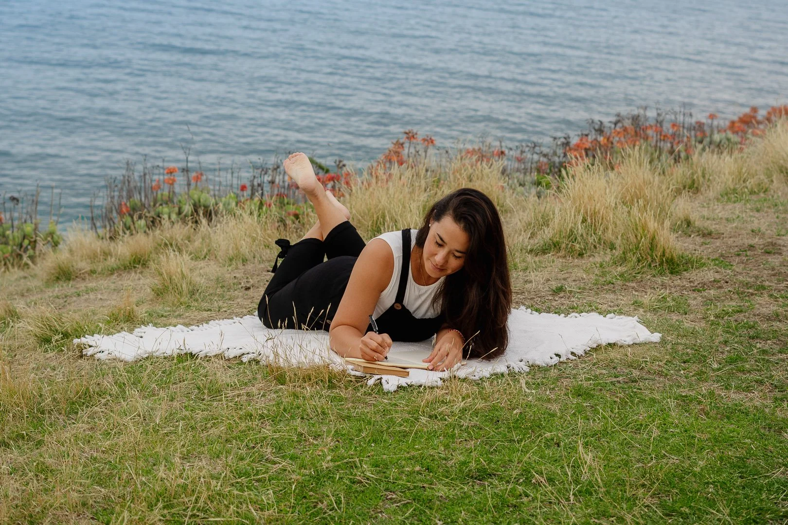 A woman lying on her stomach on a white blanket on a grassy hill near the water, writing in a notebook with a pen.