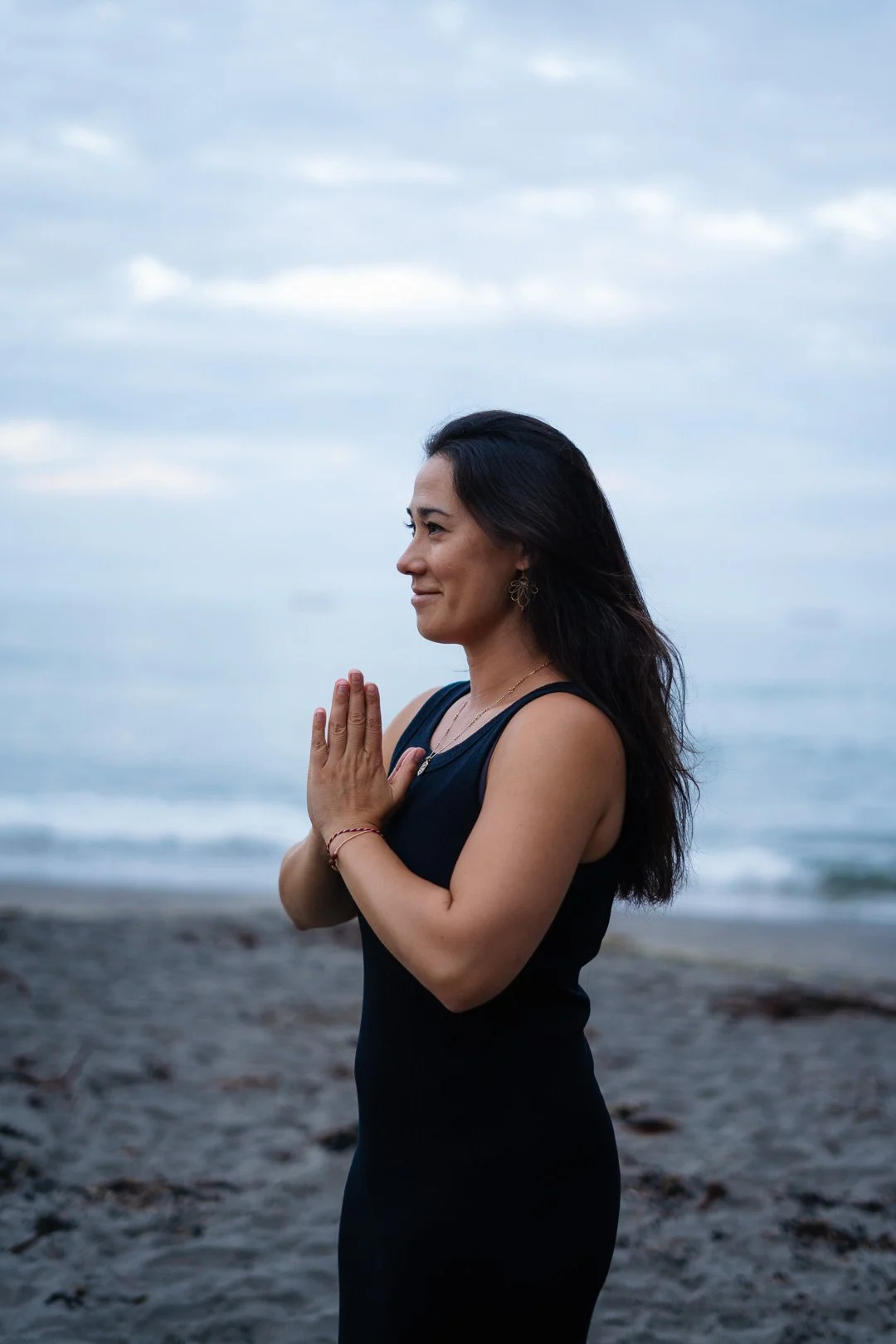 A woman standing on the beach near the ocean with her hands in a prayer position, wearing a black dress, with a calm expression, against a cloudy sky.