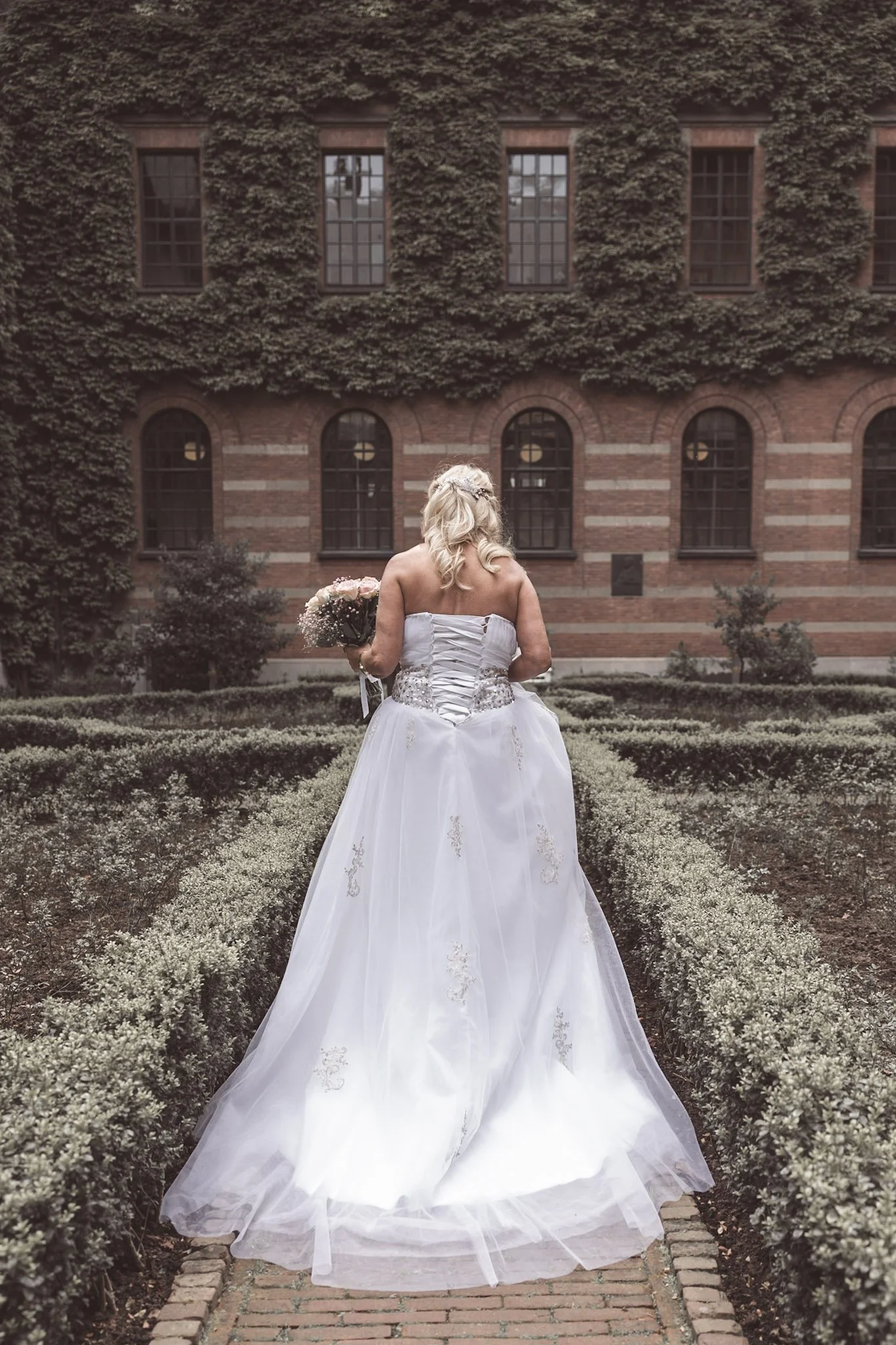 Een woman in een witte bruidsjurk staat met haar rug naar de camera in een tuin met struiken en een bloemenvaas in haar hand.