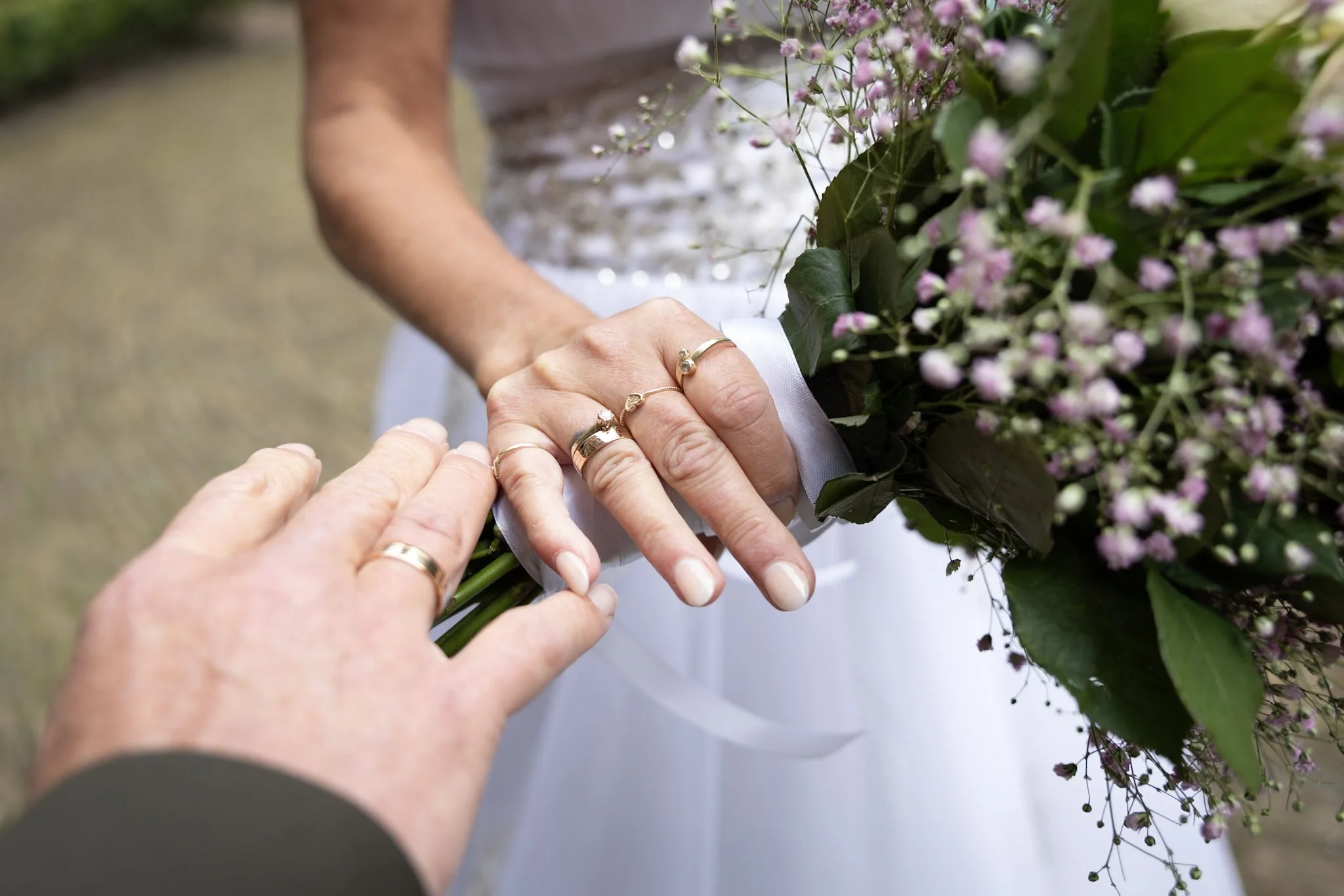 Handen die een bloemstuk vasthouden met een witte lint eromheen, terwijl een andere hand helpt bij het plaatsen van de bloem. Mensen dragen ringen en de bloem bestaat uit kleine roze en witte bloemen en grote groene bladeren.