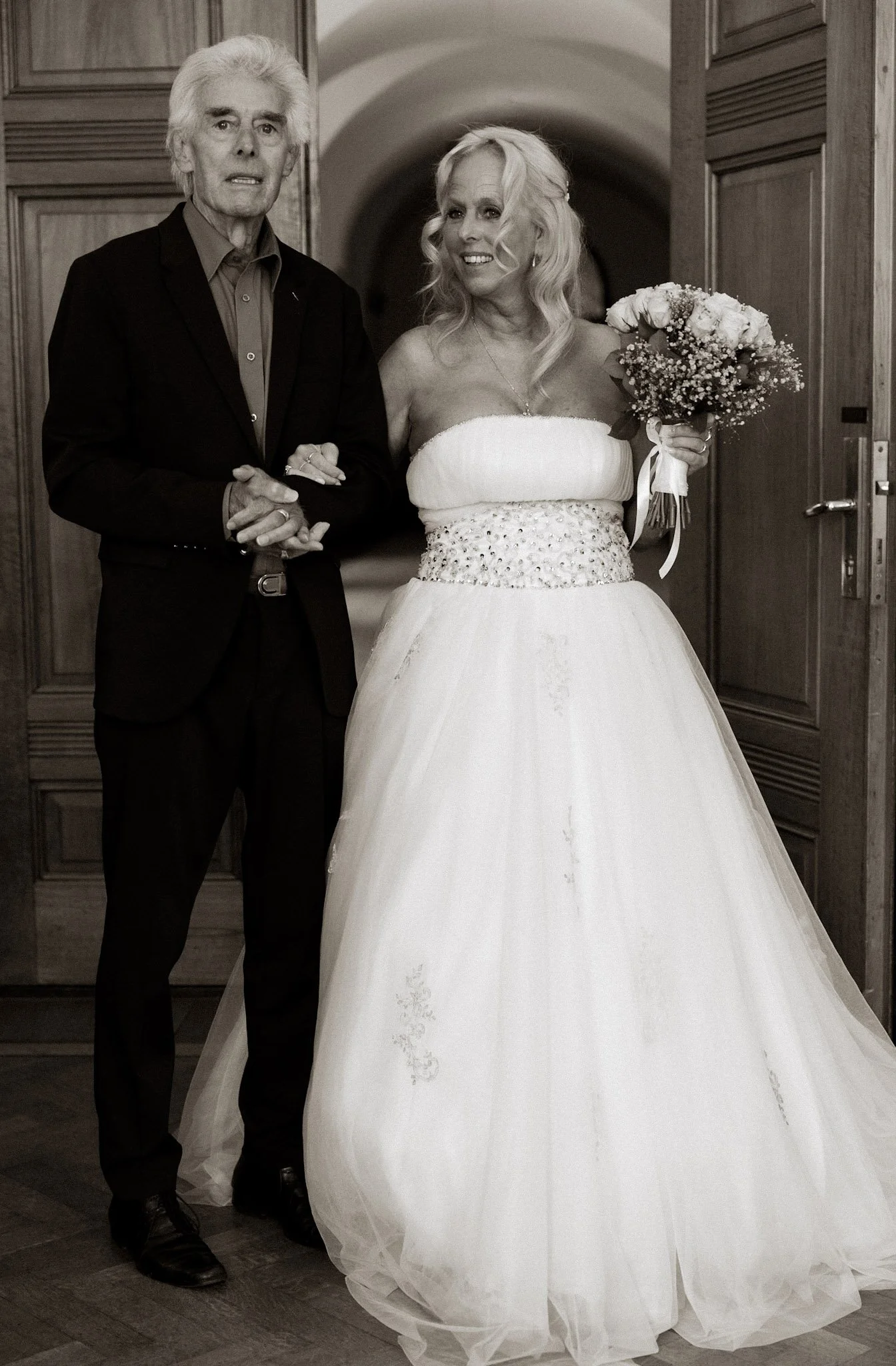 Een vrouw in een witte trouwjurk met een bouquet bloemen staat naast een man in een zwarte jas, ze houden elkaars handen vast in een hal met houten wand. De foto is zwart-wit.