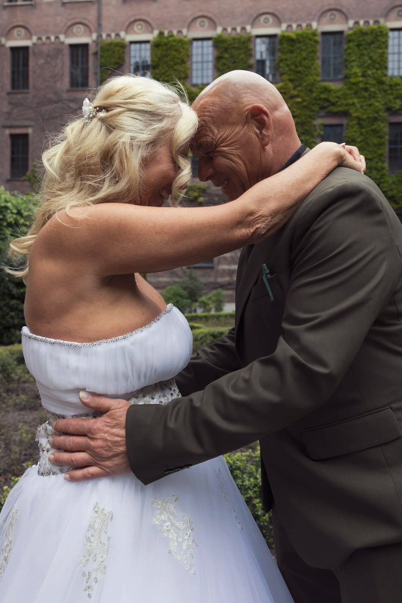Een ouderwetse bruid en bruidegom die in een tuin elkaars voorhoofd en voorarmen tegen elkaar drukken, beiden glimlachend.