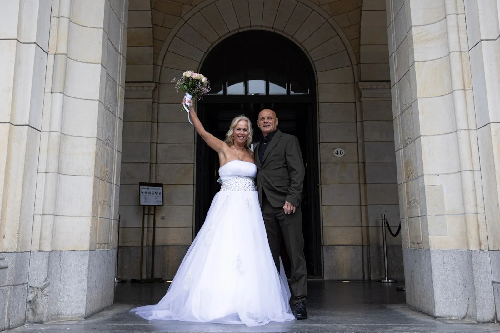Trouwfoto van een vrouw in een witte jurk met bloemen in haar hand, staande naast een man in een grijze pak voor een gebouw met een boogdeuren