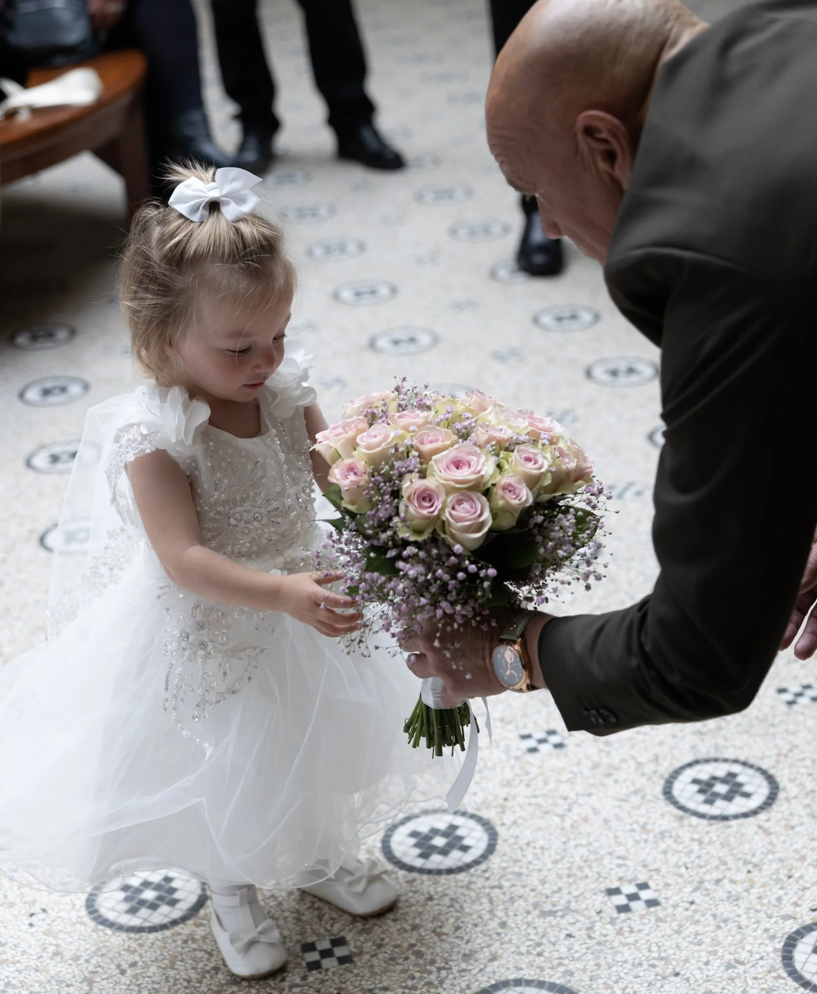 Een jonge meisje in een witte jurk met een grote witte strik in haar haar ontvangt een bos bloemen van een man tijdens een ceremonie.
