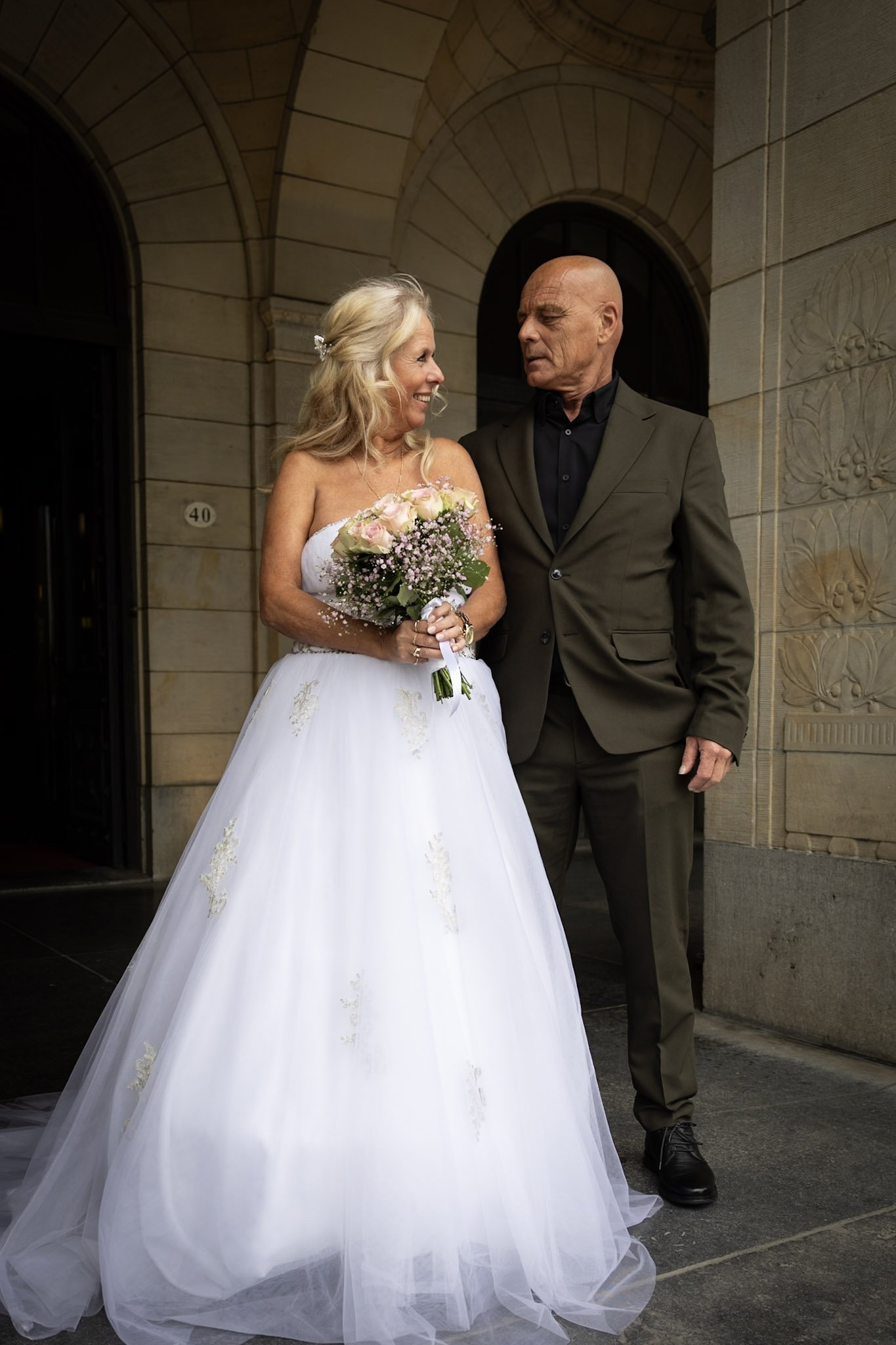 Een bruid in een witte trouwjurk met bloemen in haar haar en een man in een zwarte pak, staan voor een stenen gebouw in een formele setting.