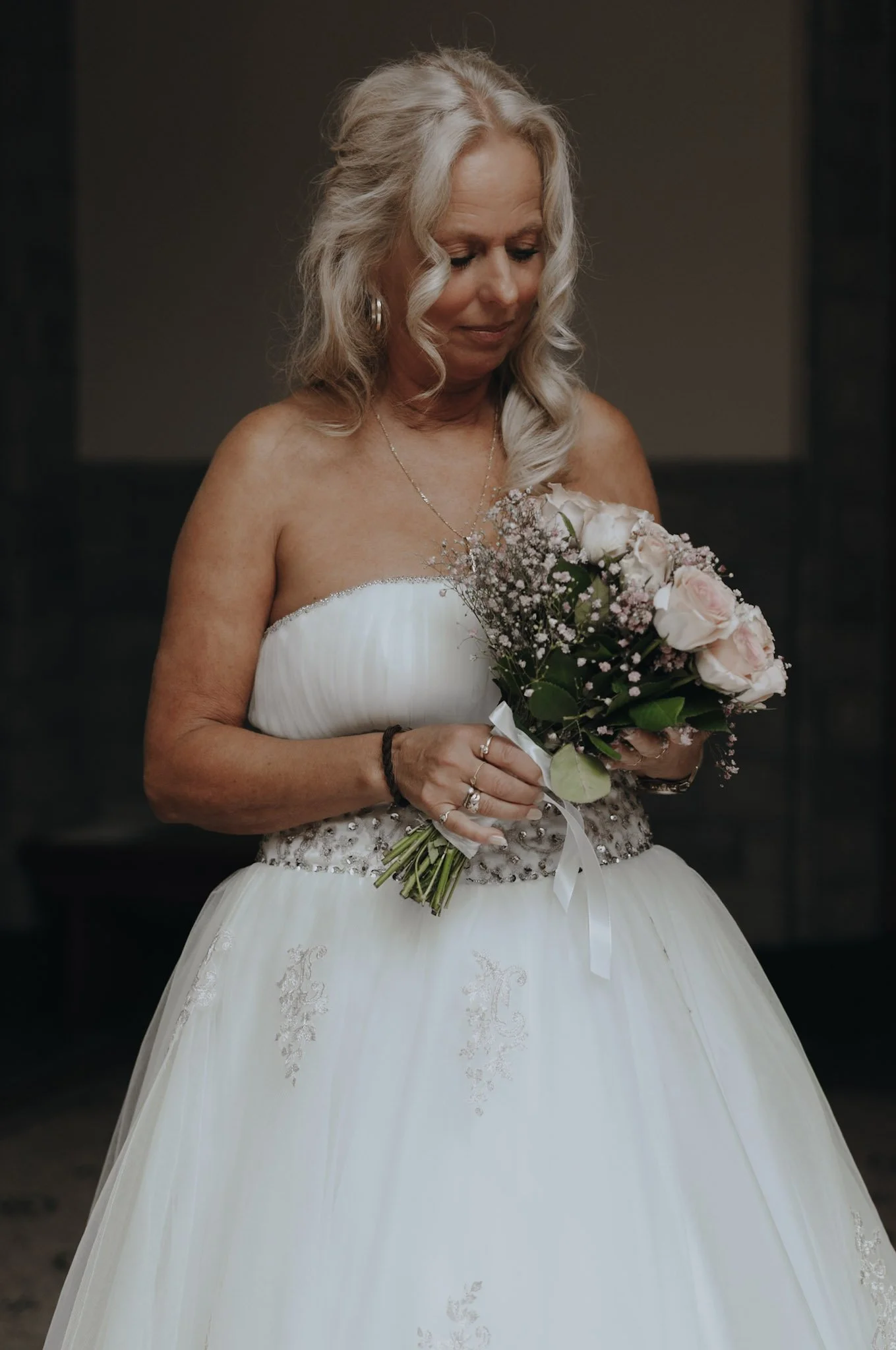 Een oudere vrouw in een witte strapless trouwjurk met een bos bloemen, haar haar gekruld en zilverachtig, glimlachend met gesloten ogen.