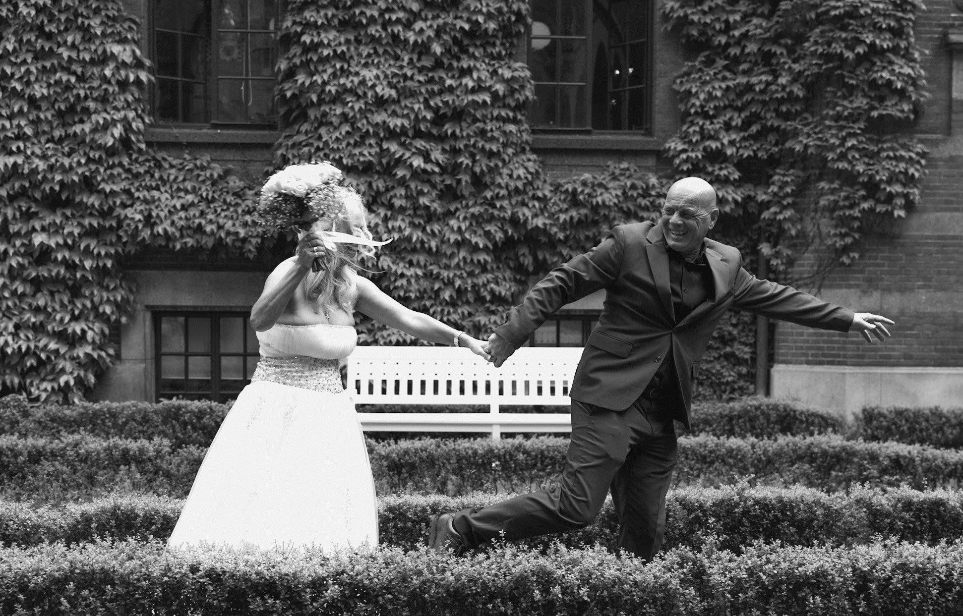 Een paar danst samen in een tuin met groene struiken en een wit bankje op de achtergrond, terwijl een vrouw een bos bloemen vasthoudt.