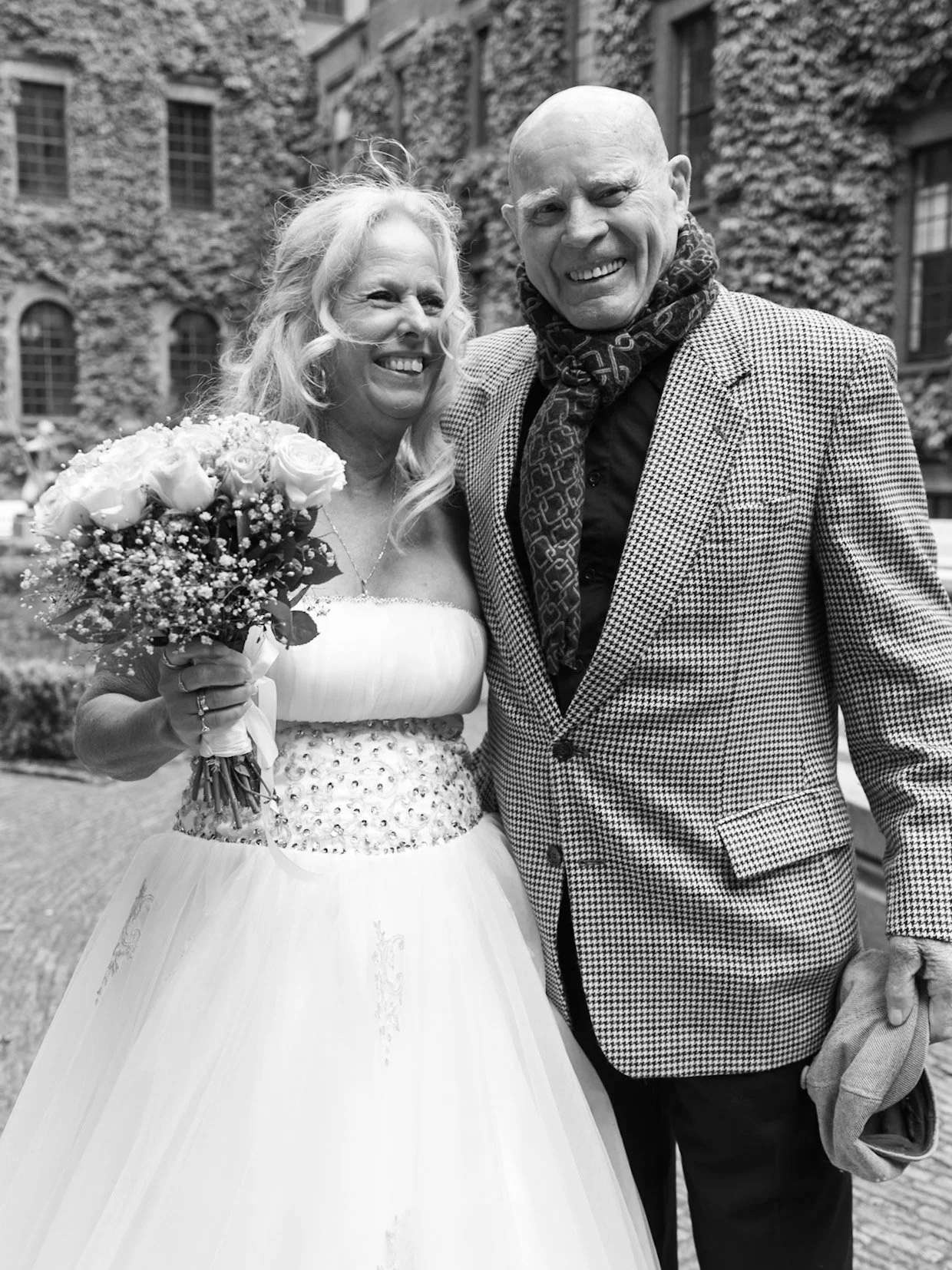 Een zwart-witfoto van een bruid en een man die bij een huwelijk poseren. De bruid glimlacht en houdt een bloemboeket vast. De man draagt een geruite jas en een sjaal.