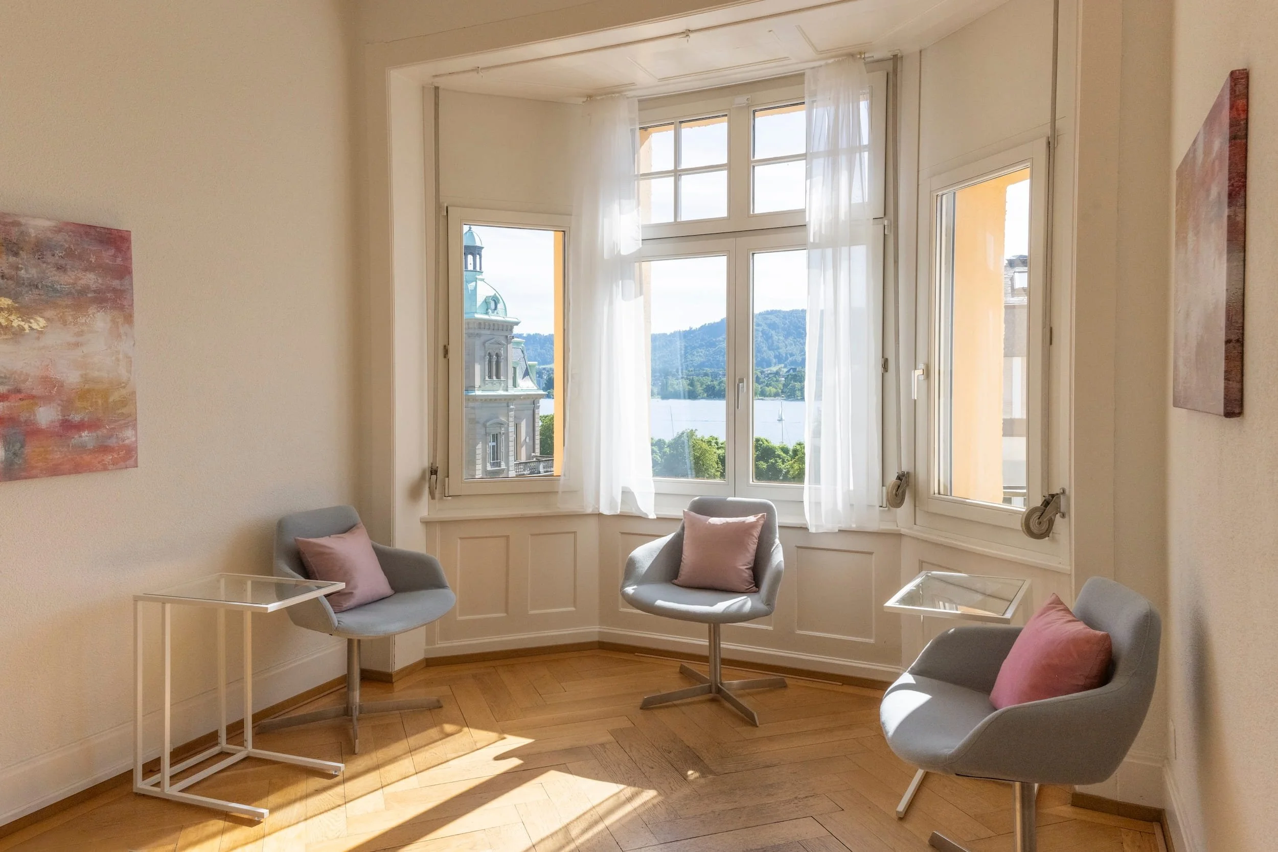 Ein Wohnzimmer mit einem großen Fenster, Vorhängen und Blick auf einen See und eine Kirche im Hintergrund. Es gibt drei graue Sessel mit Kissen und kleine Beistelltische im Raum.