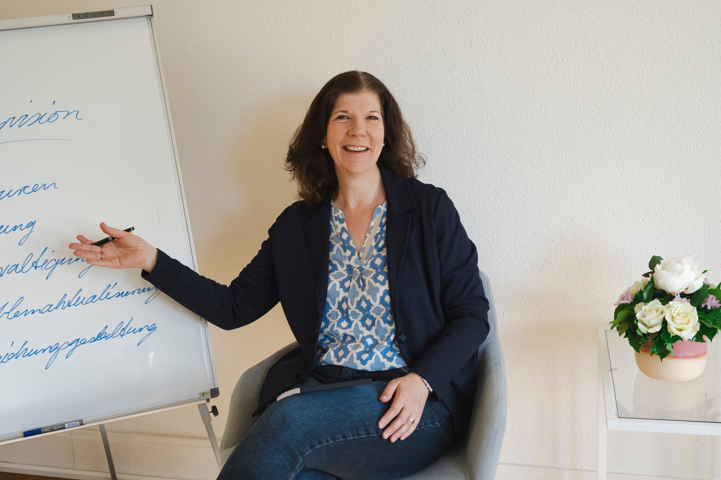 Eine lachende Frau sitzt in einem Büro neben einem Whiteboard mit blauer Handschrift. Neben ihr steht ein Topf mit weißen und pinken Blumen.