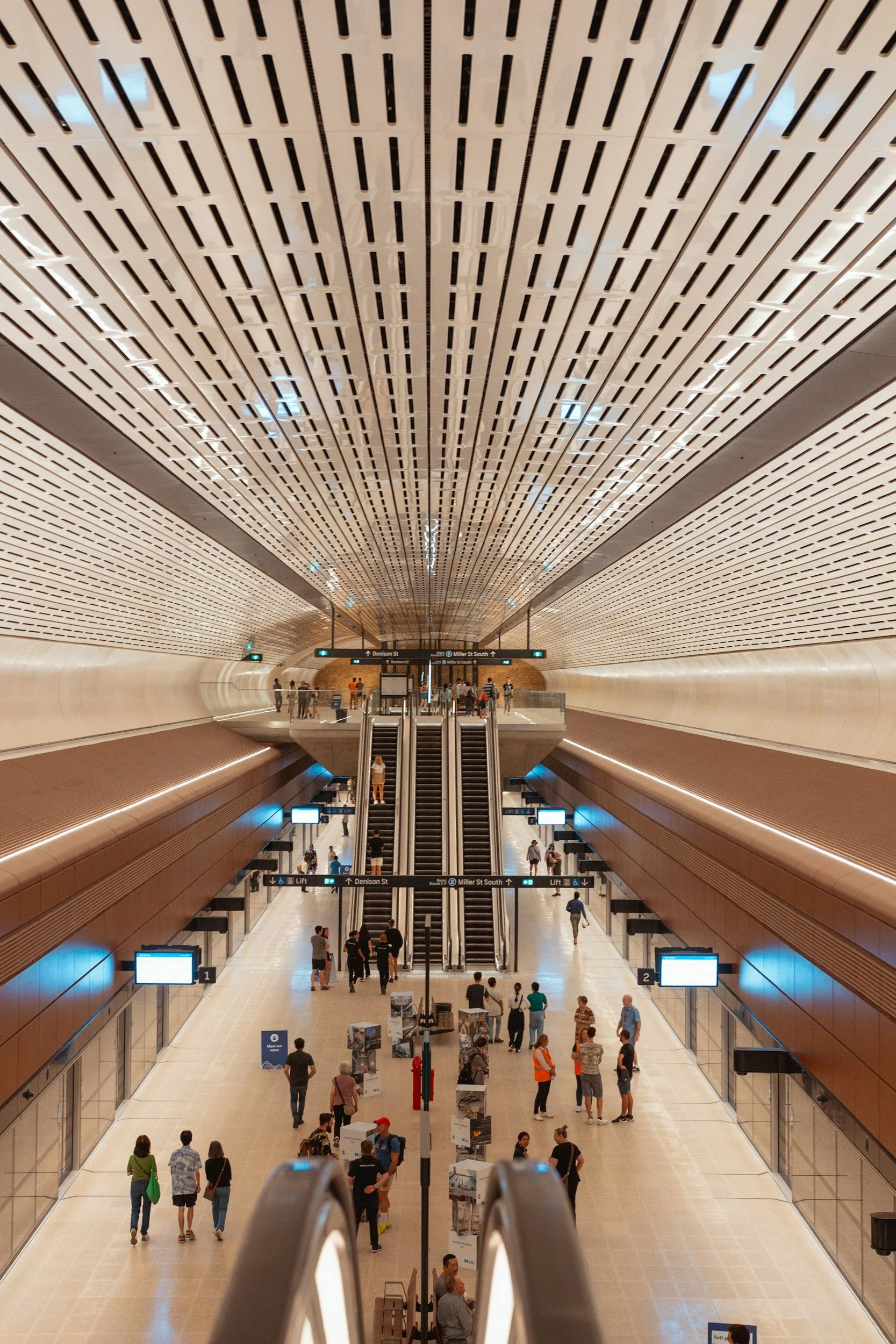 Inside a modern underground train station with a high, curved ceiling featuring a pattern of black and white geometric shapes. There are multiple levels with escalators, digital screens, and people walking or waiting.
