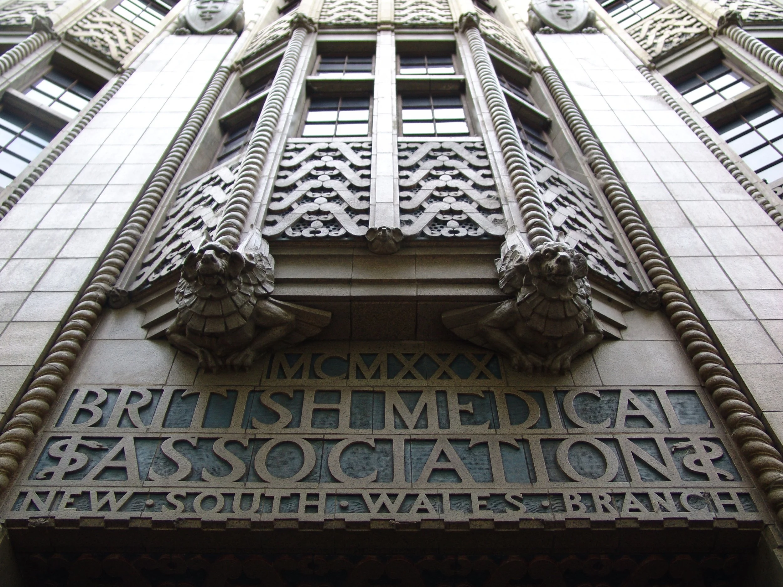 Close-up of a building facade with decorative stonework, statue lions, and engraved text, including Roman numerals and various words.