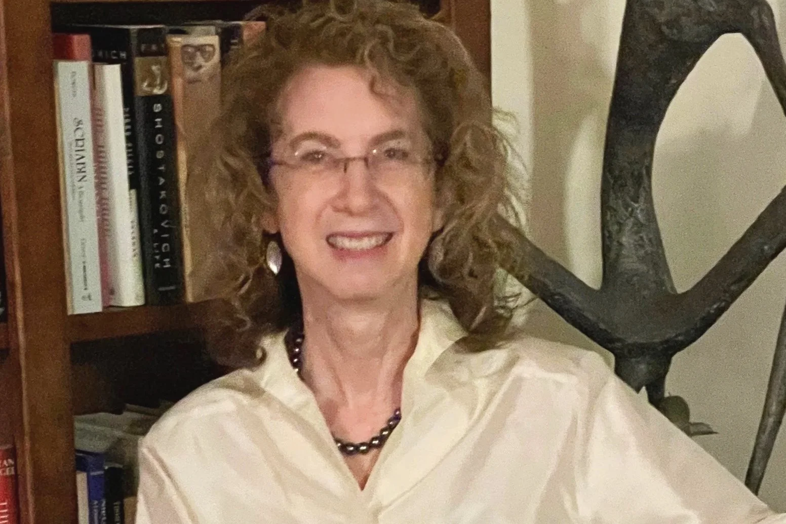 A smiling woman with curly, shoulder-length hair wearing glasses, a light-colored blouse, a dark beaded necklace, and earrings. She is sitting in front of a wooden bookshelf filled with books and in front of a dark decorative wrought-iron object.