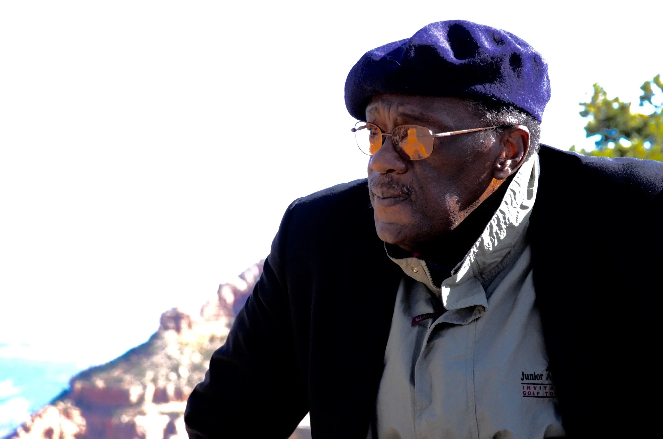 An elderly man wearing glasses, a purple beret, and a black jacket outdoors with a bright sky and trees in the background.