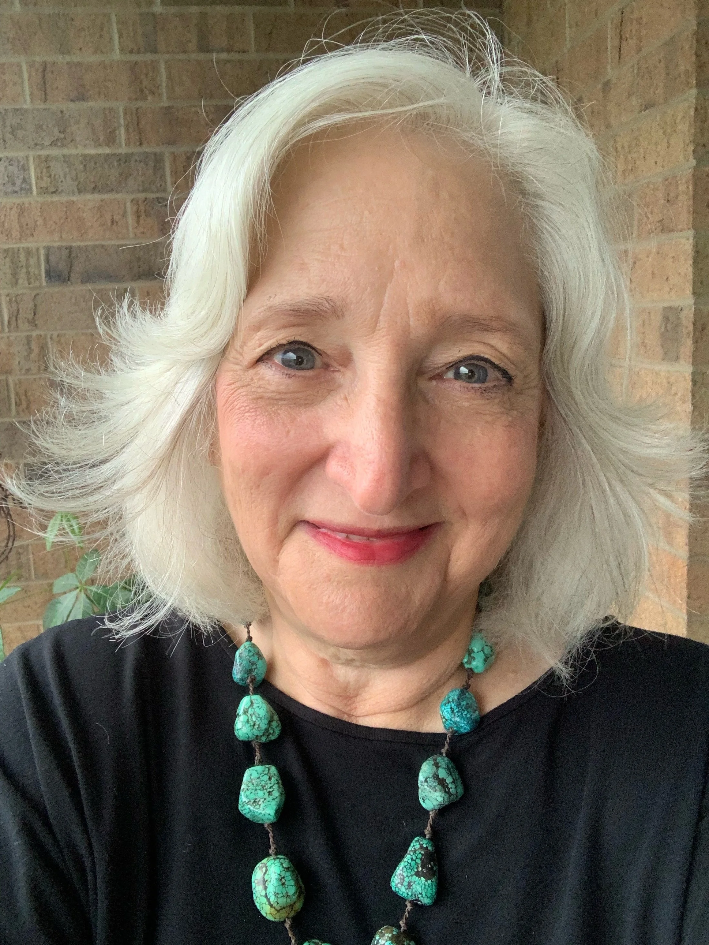 Close-up of an older woman with white hair styled in loose waves, wearing a black top and a turquoise beaded necklace with matching earrings, standing against a brick wall.