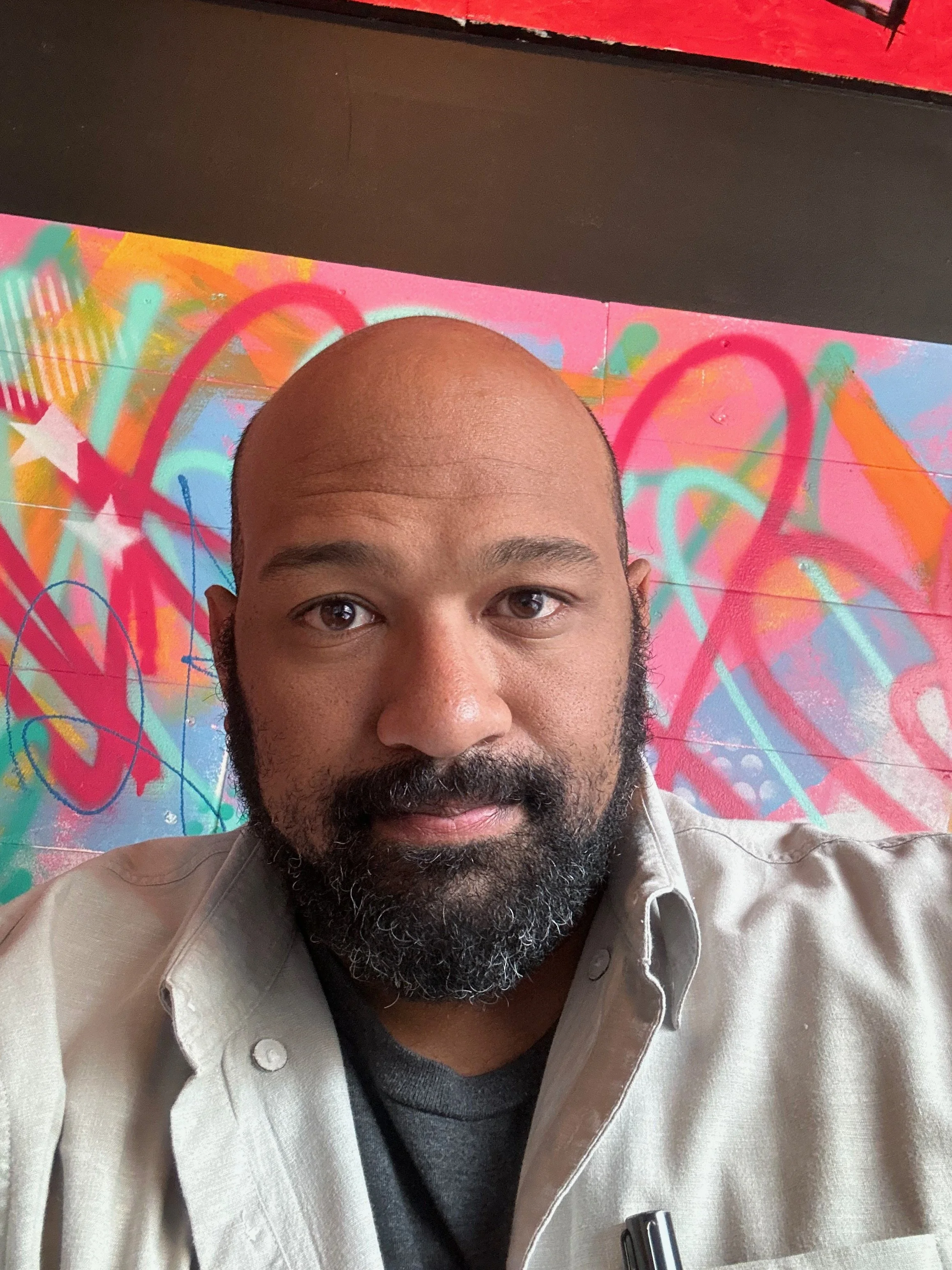 A man with dark skin, a beard, and a mustache, wearing a beige jacket over a dark shirt, taking a selfie in front of a colorful graffiti art wall with pink, orange, green, and blue abstract designs.