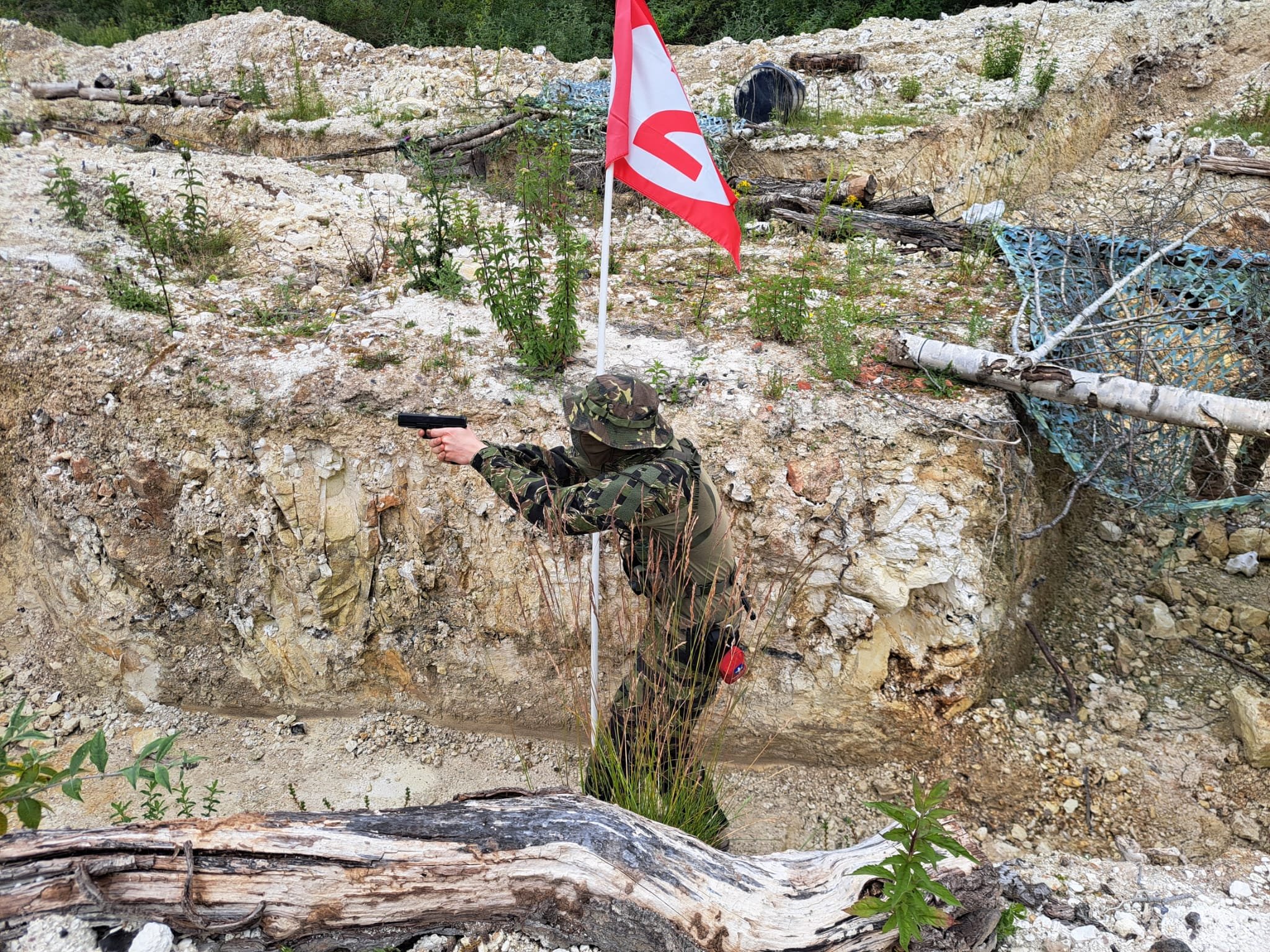 A dirt and gravel hillside with scattered debris, logs, and plants. A security or warning flag is anchored in the ground near the center, with a person in camouflage clothing holding a handgun walking in the lower part of the image.