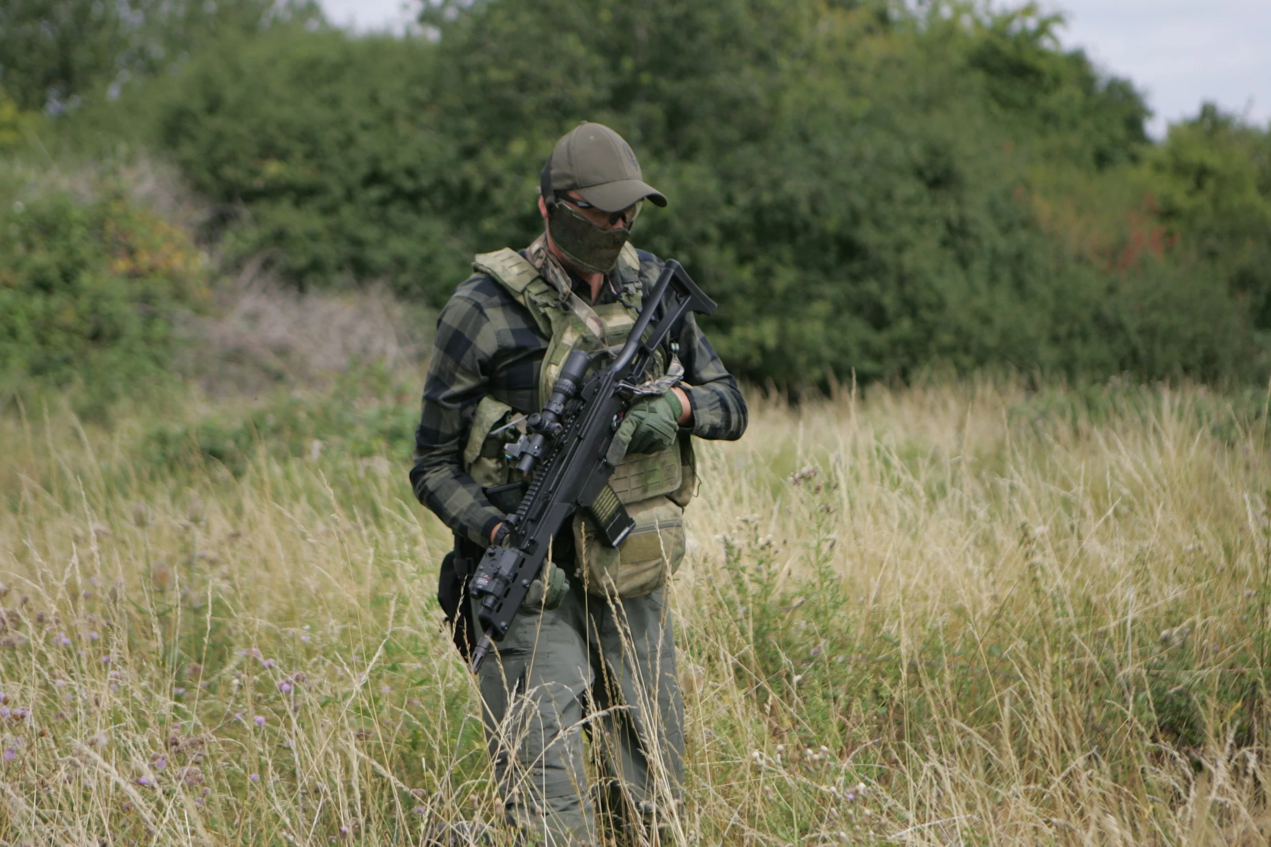 A person dressed in military camouflage gear with a helmet, sunglasses, and face covering, holding a rifle while walking through a grassy field with trees in the background.