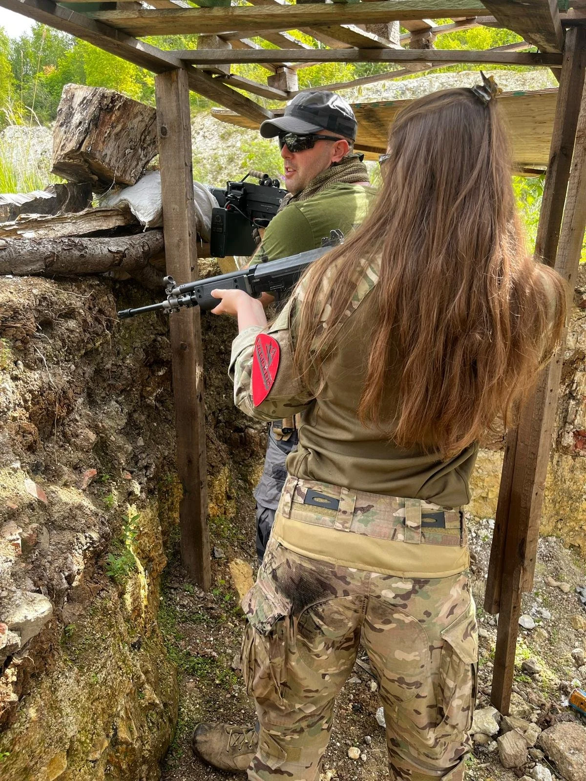 Two individuals in military-style camouflage clothing aiming rifles from a trench with a wooden barricade, surrounded by trees and natural terrain.