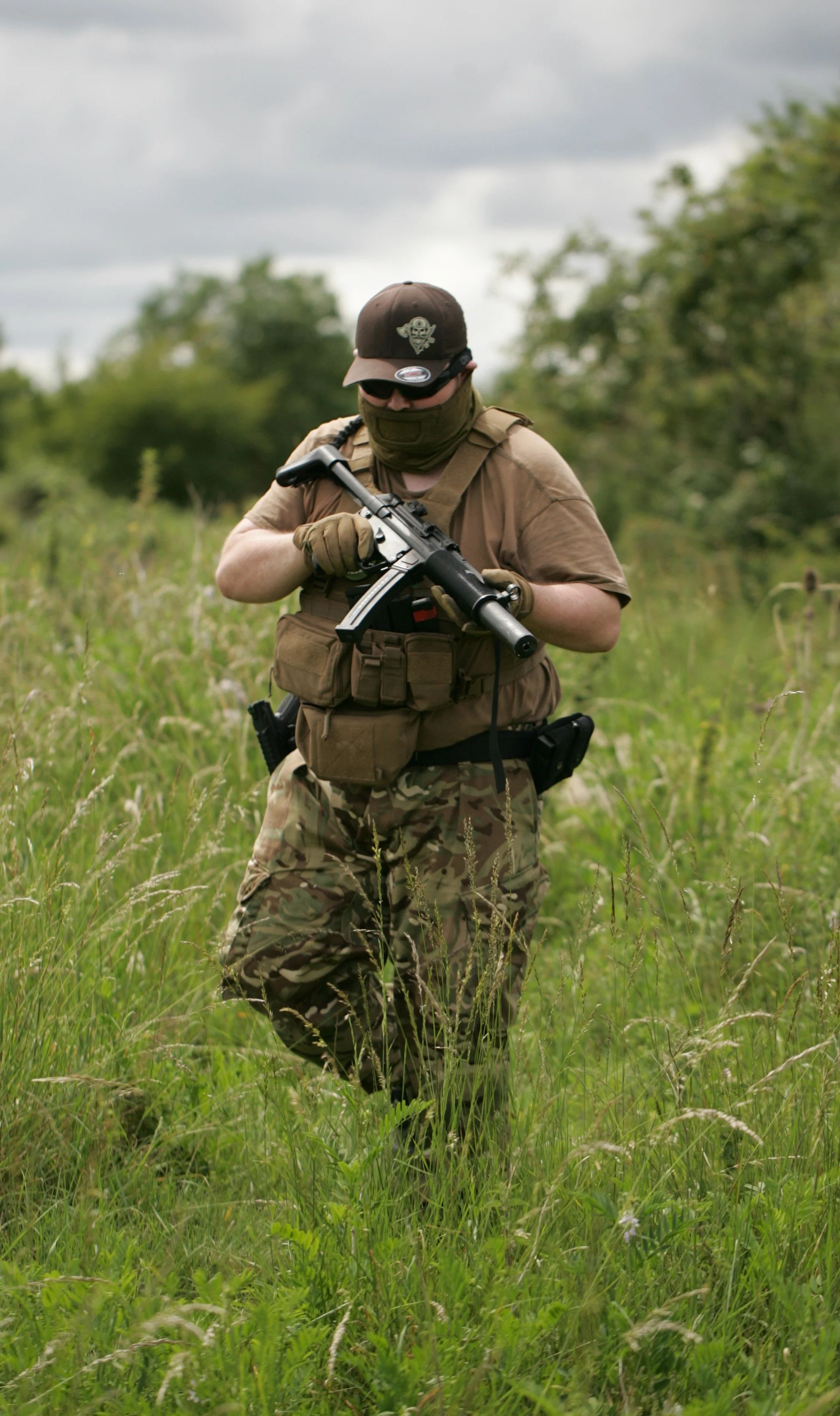 A person dressed in camouflage pants, tan shirt, tactical vest, black cap with skull emblem, sunglasses, and face covering, walking through tall grass with trees and cloudy sky in the background, holding a firearm.