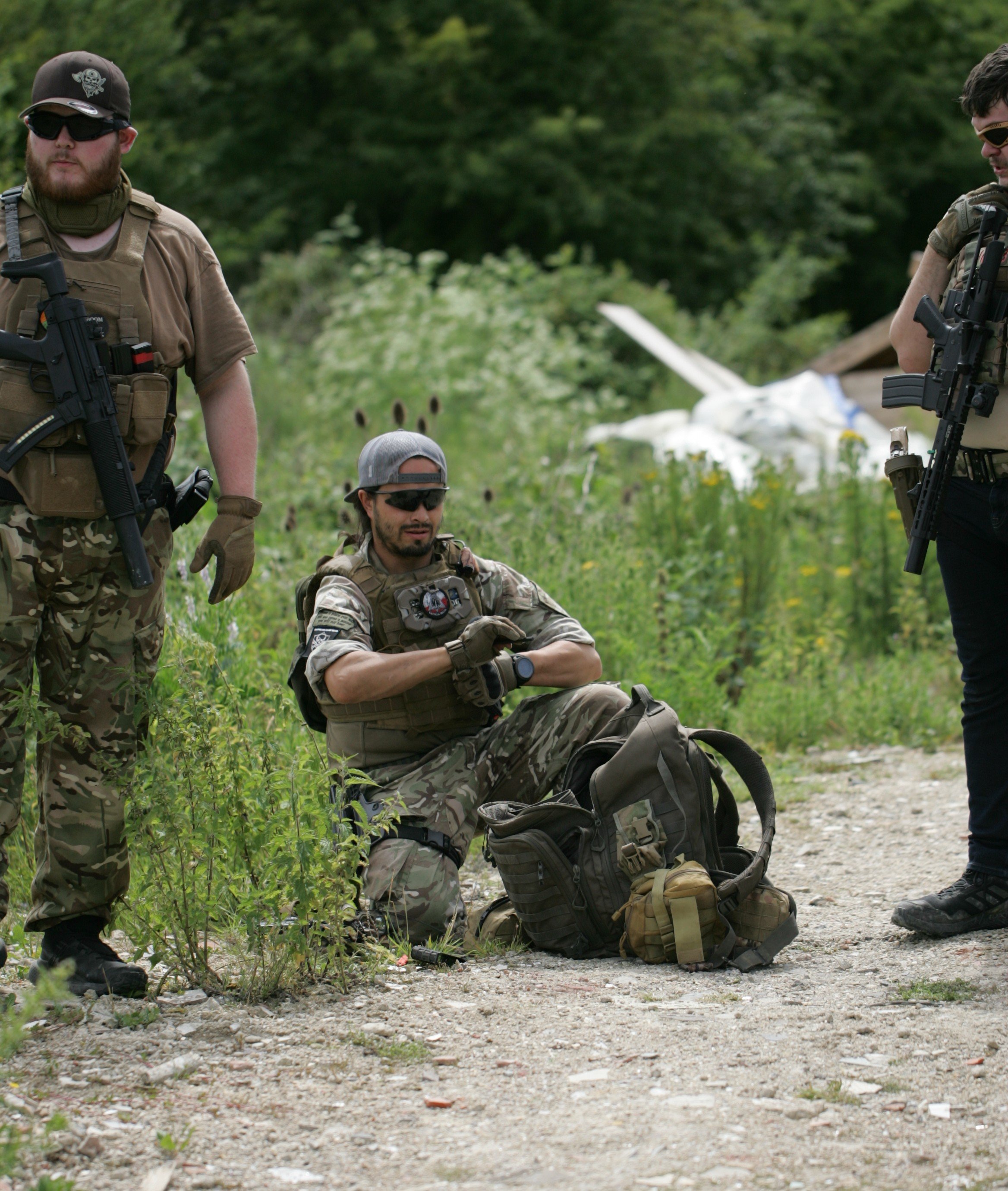 Three soldiers in camouflage and tactical gear resting outdoors in a green, wooded area. One soldier with a beard and sunglasses stands on the left, a soldier with tattoos and sunglasses sits in the middle, and a third soldier stands on the right wit