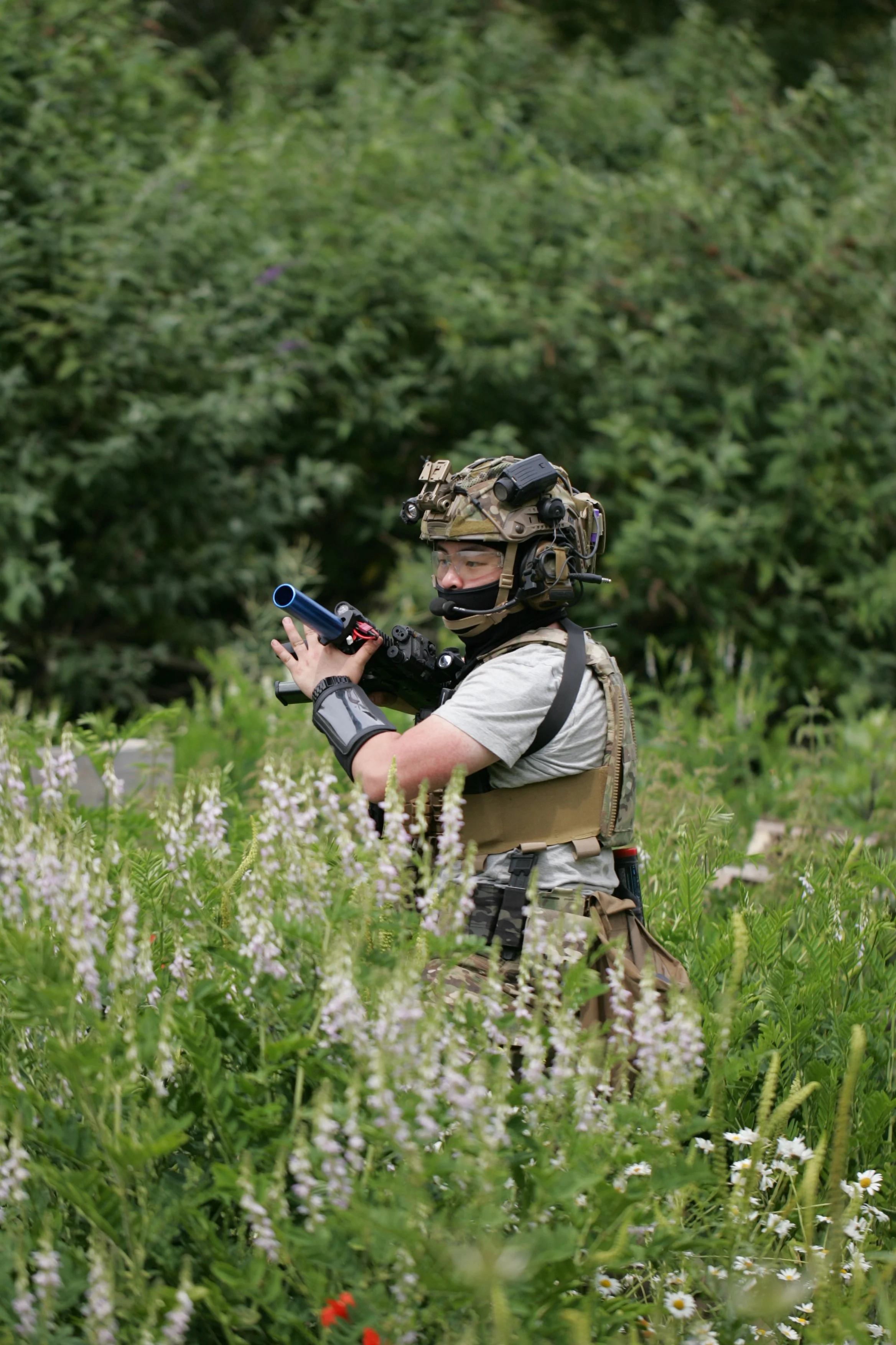A soldier in camouflage gear and tactical vest holding a firearm with a scope, walking through a lush, green, flowering field with trees in the background.
