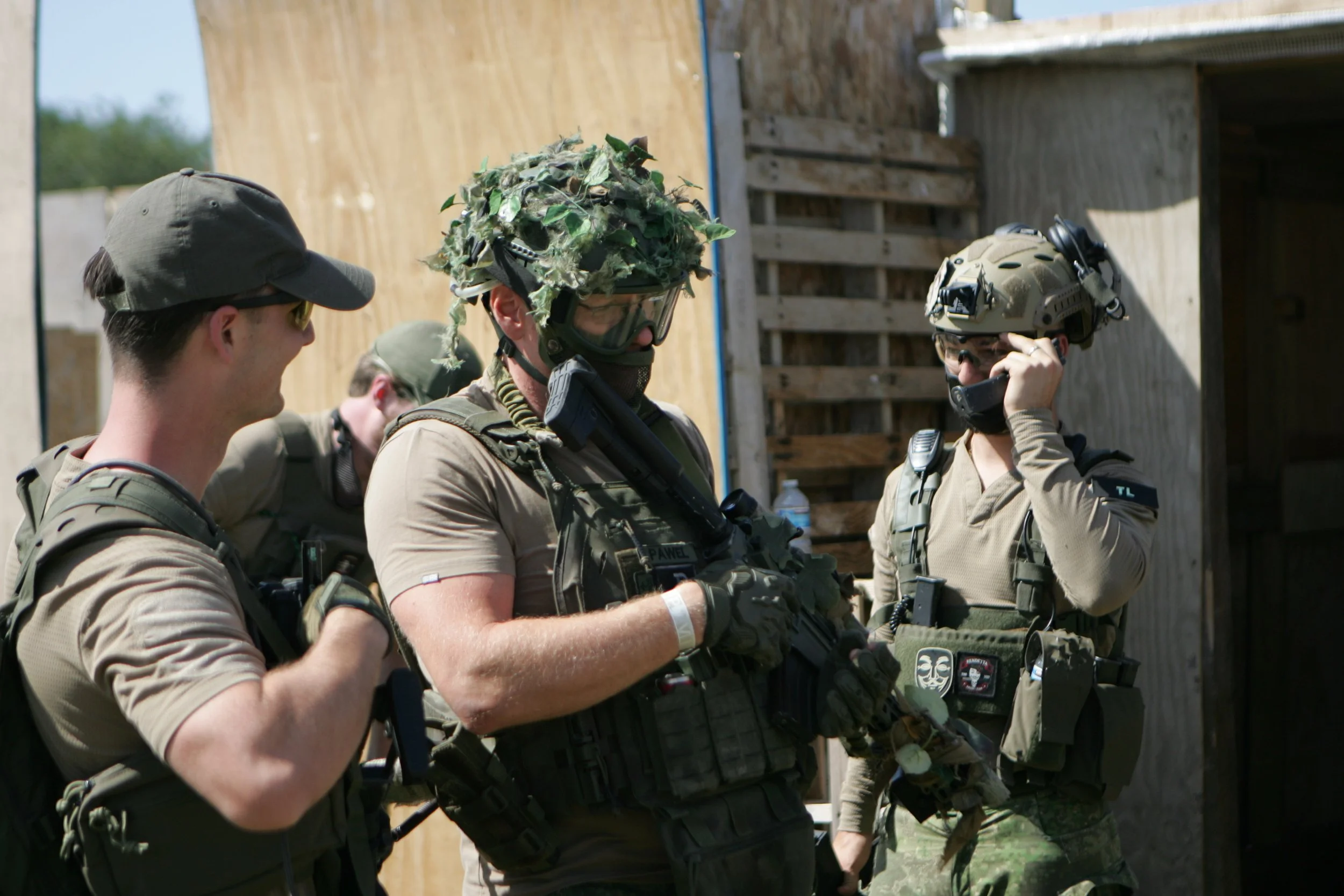 Military soldiers in uniform and tactical gear, one wearing a camouflage helmet with leaves, engaging in a training Exercise outdoors near a wooden structure.