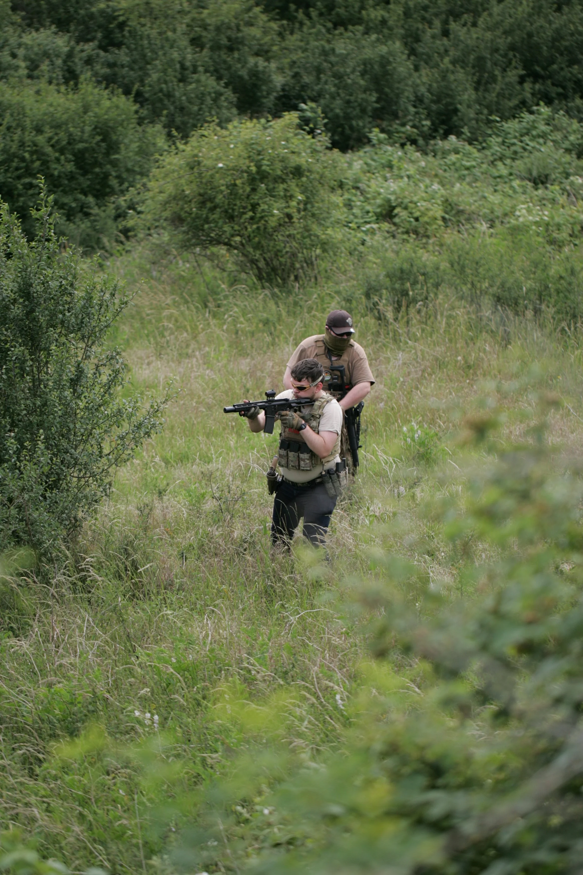 Two individuals in tactical gear walking through grassy, wooded area, one aiming a rifle.